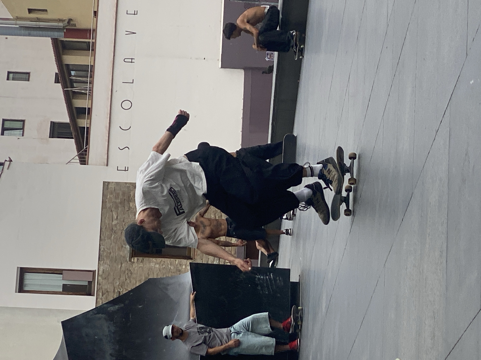This daytime photograph captures an active skateboarding scene in an urban environment in Barcelona, Spain. The setting appears to be a public square or a skatepark, characterized by various concrete structures and surrounding buildings.
In the central foreground, a male skateboarder is depicted mid-trick, appearing to be airborne or in the process of landing. He wears a white t-shirt, loose black trousers, and a black cap. A black wrist brace is visible on his left arm, which is bent at the elbow. His feet, clad in light-colored sneakers with white socks, are positioned on his skateboard.
Several other individuals are present. To the lower left, a person wearing a gray t-shirt, light blue shorts, and a white cap sits casually on a large, angular black structure, holding its edge. Behind the main skater, another person in black clothing is partially visible. Further back, near the black structure, a shirtless individual bends over. In the upper right background, on an elevated concrete platform, another shirtless person interacts with a skateboard.
The ground is a light grey concrete surface, marked with linear patterns. The background features multi-story urban buildings. A prominent white building on the left has several dark-framed windows and a ladder extending upwards. Vertically displayed on this white building are the block letters "ESCOLAVE". The lighting suggests bright, possibly overcast, daytime conditions with no harsh shadows. The overall atmosphere is one of casual activity and urban recreation.