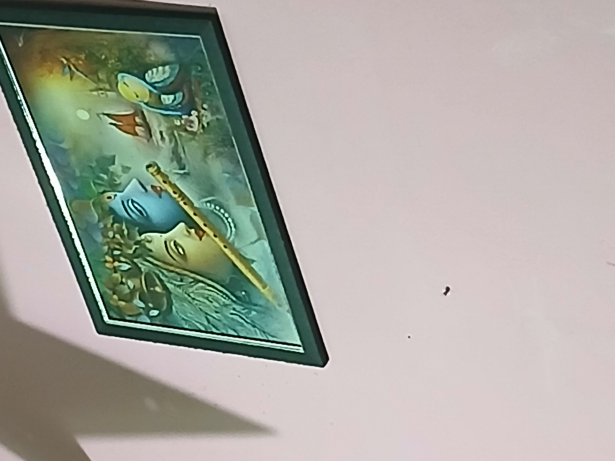 A framed devotional painting is prominently displayed on a light pink wall, captured from an upward, diagonal angle. The painting depicts a figure, likely Lord Krishna, with light blue skin and a serene expression, eyes gently closed or downcast. A golden flute is held diagonally across the mid-section of the artwork, resting near the figure's face.
Above the main figure, two distinct birds are visible: one larger, stylized bird with yellow and blue plumage, and a smaller bird with reddish-orange wings. A luminous, circular orb, possibly a moon or celestial body, emanates light from the upper left corner of the painting. The background features soft, blended colors suggestive of foliage or clouds, primarily in shades of green and blue.
The artwork is enclosed in a dark, possibly black, frame with a thin, reflective inner border. A strong, angular shadow is cast by the frame onto the light pink wall, indicating an off-camera light source. Two small, dark specks are visible on the bare wall to the right of the frame. The overall scene suggests an indoor setting, possibly a home or temple. There is no visible text in the image.