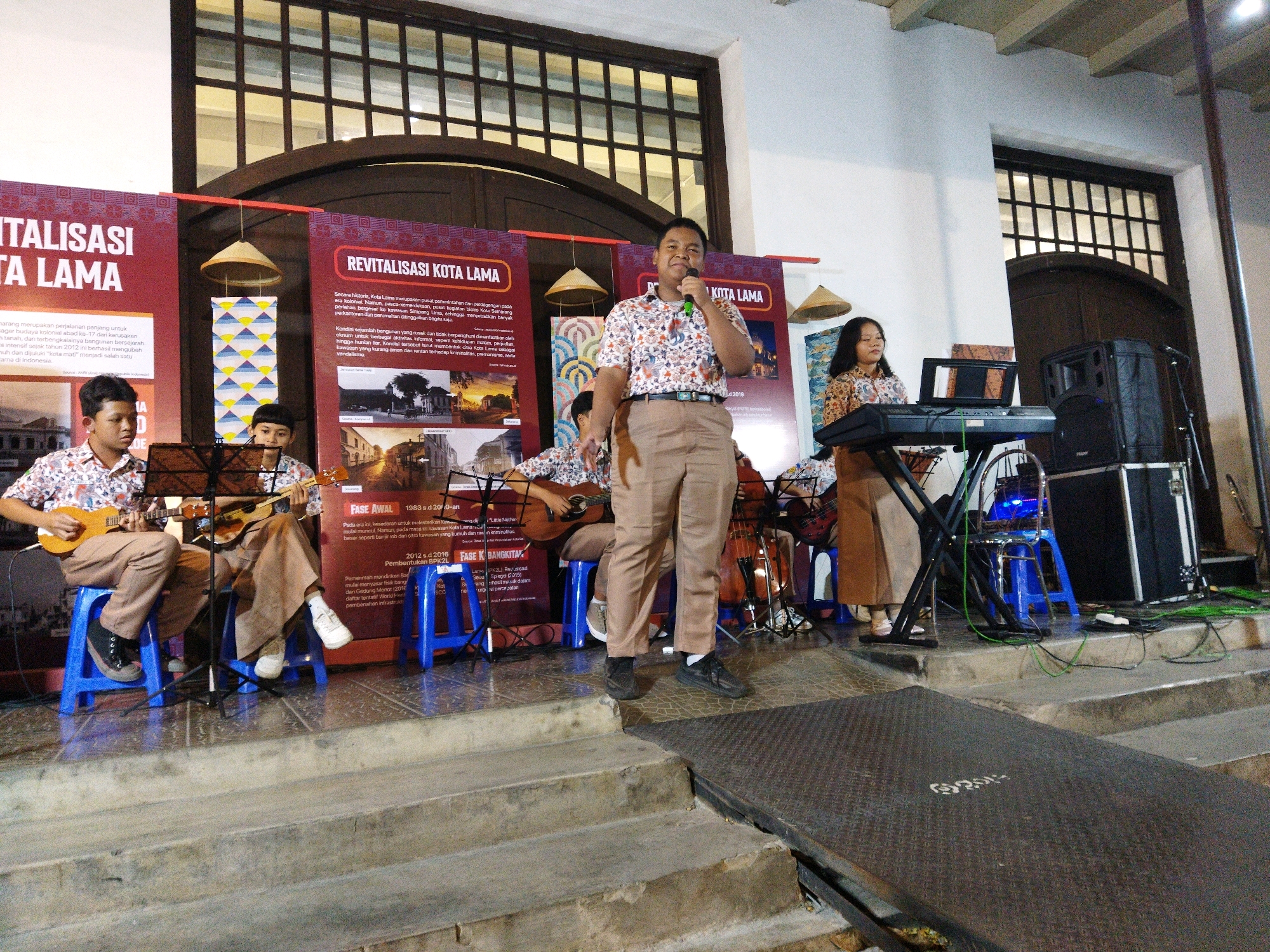 The image depicts an outdoor event at Kota Lama, Semarang, Indonesia, likely a performance or presentation. Four young people are on a raised stage. In the foreground, two boys sit on blue plastic stools, each playing a ukulele. Behind them, two other individuals are also performing; one is standing and speaking into a microphone, while the other sits and plays a keyboard. The standing performer wears a floral-patterned shirt and khaki pants, and the keyboard player wears a similar patterned shirt and a brown skirt. The stage is decorated with banners that read "REVITALISASI KOTA LAMA" (Revitalization of the Old City). The background shows architectural elements typical of Kota Lama, including arched windows and weathered facades. The lighting suggests it is late afternoon or early evening. A speaker system and musical equipment are visible on the right side of the stage.