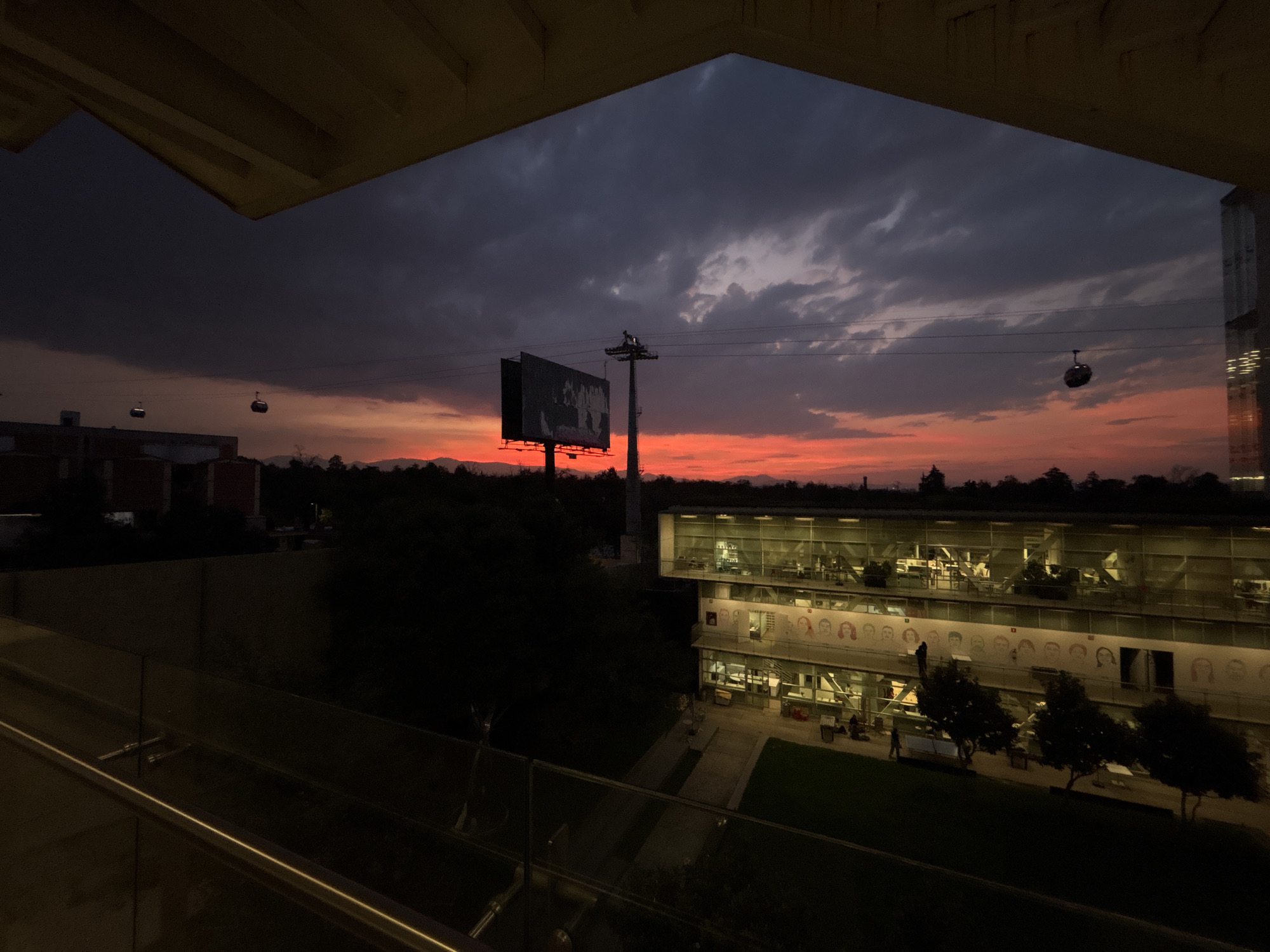 The image shows a cityscape at dusk, likely in Mexico City based on the cable cars visible in the scene. The view is framed by an architectural structure, possibly a balcony or part of a building. A large billboard and a tower stand prominently against the backdrop of the sky, which transitions from a dark, cloudy gray to a vibrant orange-red near the horizon. To the right, a modern building with glass walls and multiple stories is lit from within, revealing interior spaces and activity. A row of faces is visible on the building's facade. The foreground is dark, with a railing visible along the bottom edge of the frame. The environment includes trees and other greenery, blending into the landscape below. Cable cars are visible on the sky. People are observed on the outside of the modern glass building, walking on the balcony in front of the building, along with various objects such as benches and trees.