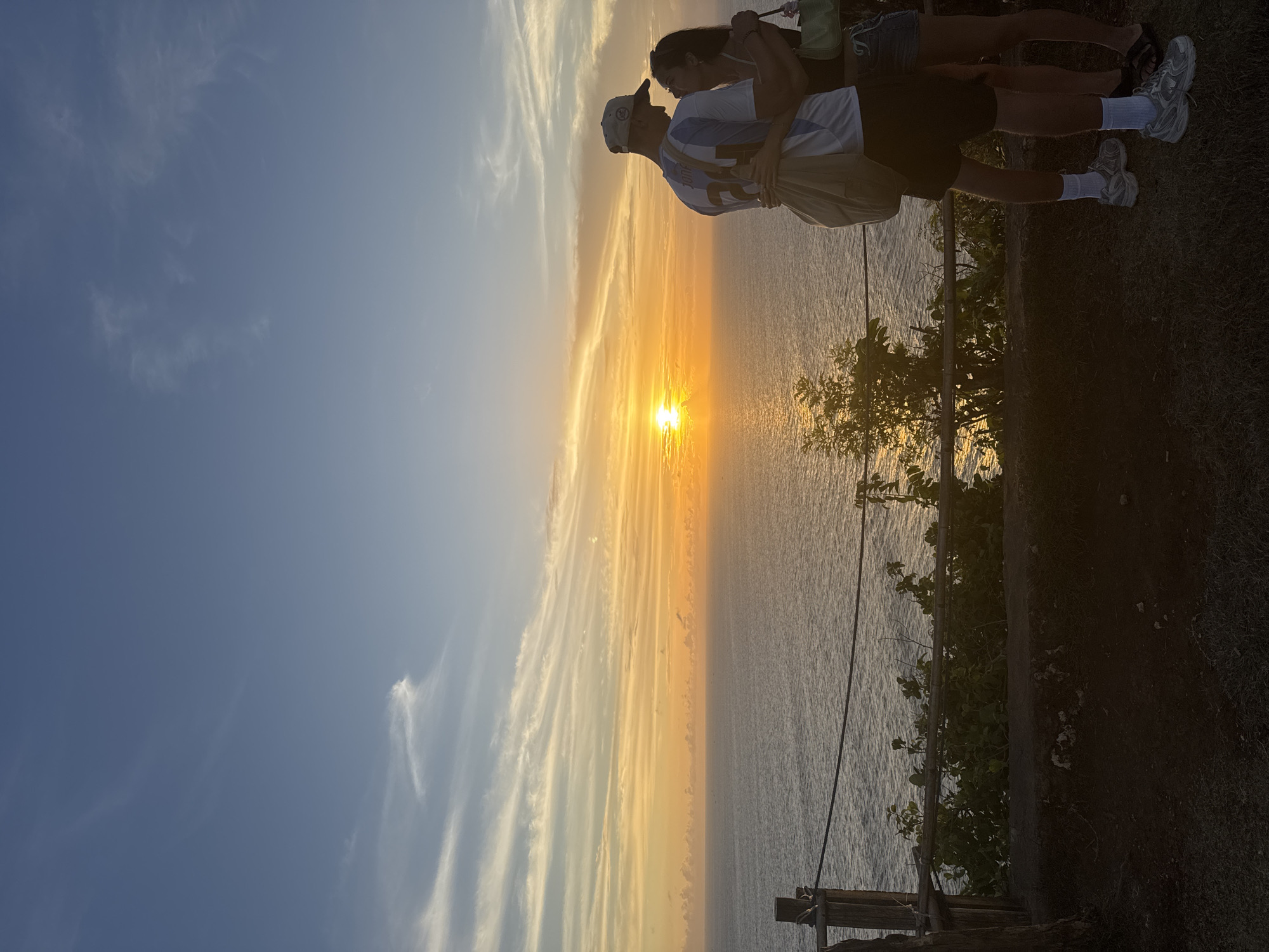 A photograph shows two people standing on a grassy area overlooking the sea during sunset. The couple is embracing, with the person on the left wearing a white and blue soccer jersey and a baseball cap. To the right of the couple is vegetation. In the background, the sun is setting over the water, creating a gradient of orange, yellow, and blue in the sky. Wooden posts and a rope are visible along the edge of the cliff.