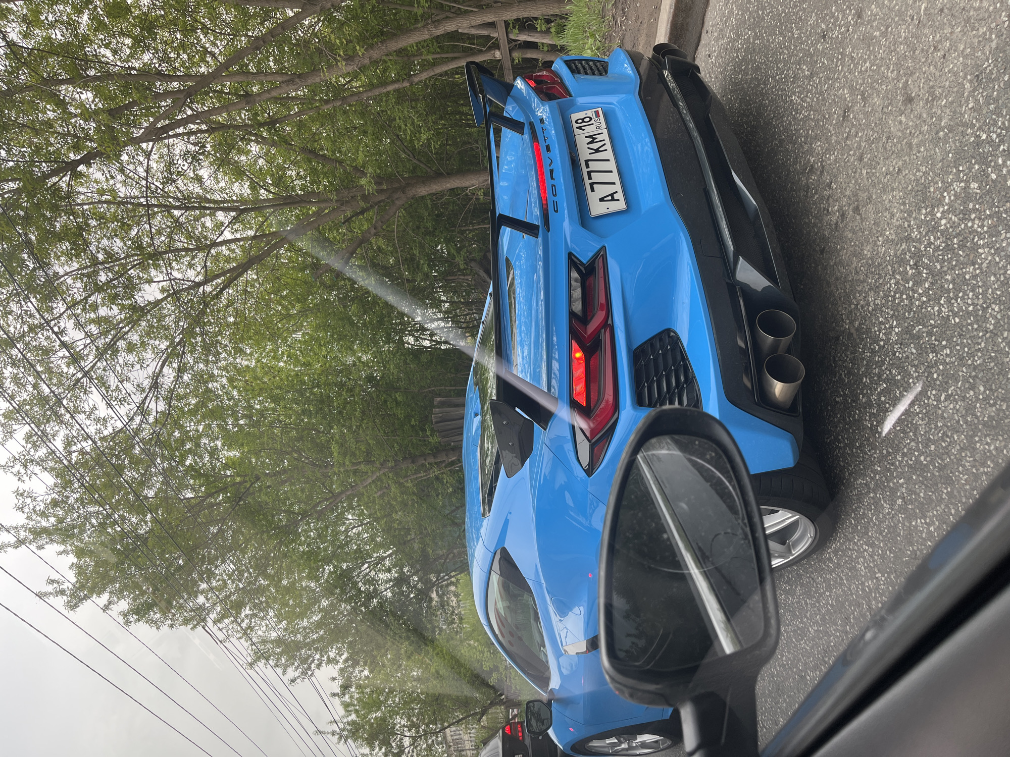 A blue Corvette with license plate A777KM18, registered in Russia, is visible from the side mirror of another car. The location is likely in Izhevsk, Russia based on the license plate. The car is on a paved road surrounded by trees.