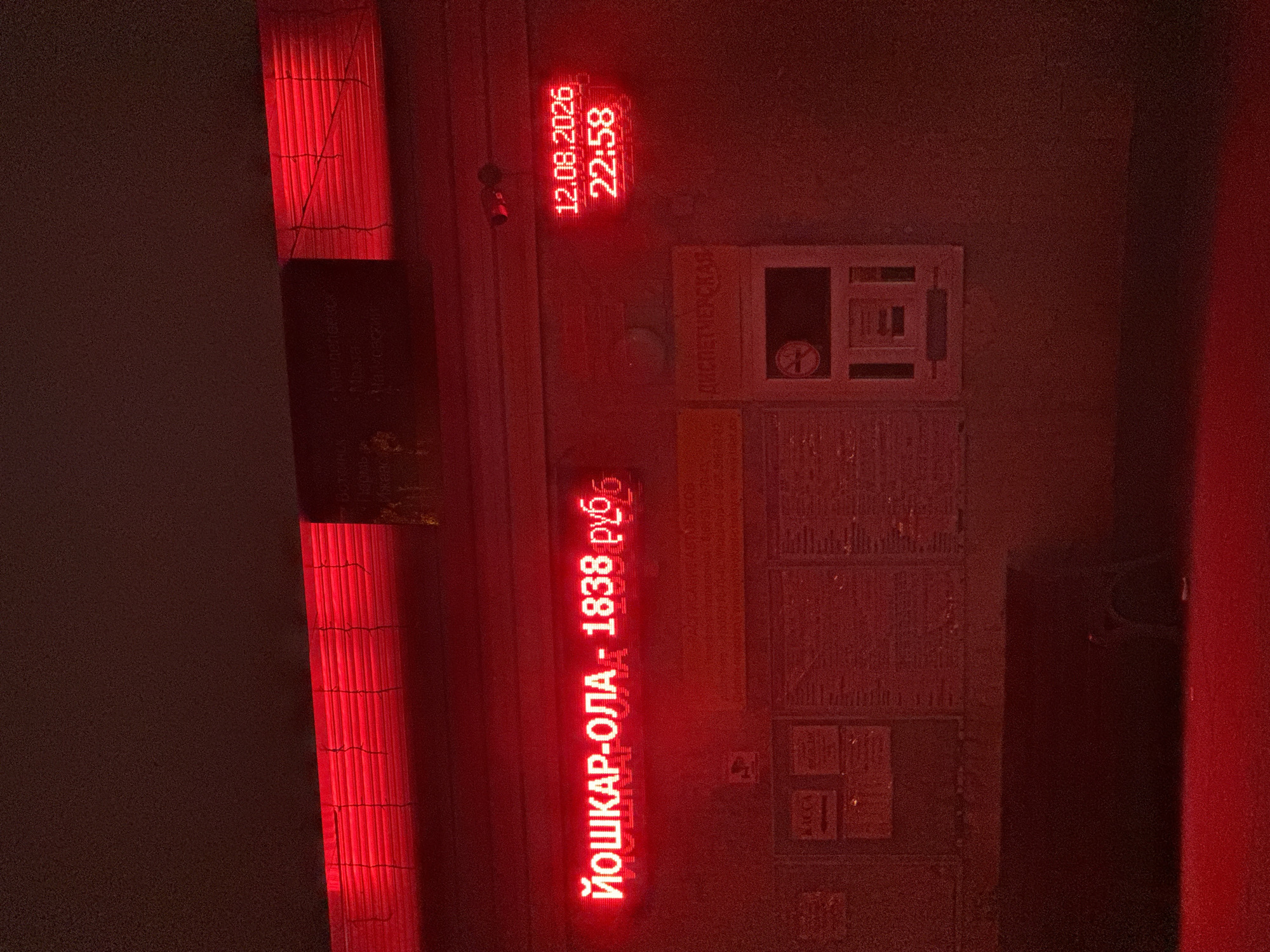 The image shows a nighttime scene illuminated by red neon lights. The primary focus is a bright red LED display that reads "ЙОШКАР-ОЛА - 1838 руб." This translates to "Yoshkar-Ola - 1838 rubles." To the right of this, another red LED display shows the date "12.08.2026" and the time "22:58".
Above the main LED display, a dark horizontal structure with red lighting behind a set of blinds is visible. Below the displays, there are several posters or information boards. One poster on the left has a red graphic, and another poster to the right contains text and a graphic of a cigarette with a diagonal line through it, indicating a no-smoking area.
The overall environment appears to be an outdoor urban setting, possibly a bus station or a similar public transport point, given the prominent destination and price display, and the information boards. The red lighting creates a stark and somewhat dramatic atmosphere. There are no people visible in the image. The ground in the foreground is also illuminated by the red light, suggesting it's a paved surface. The city of Staroe Abdulovo, Russia is not explicitly mentioned or visually depicted in the image.