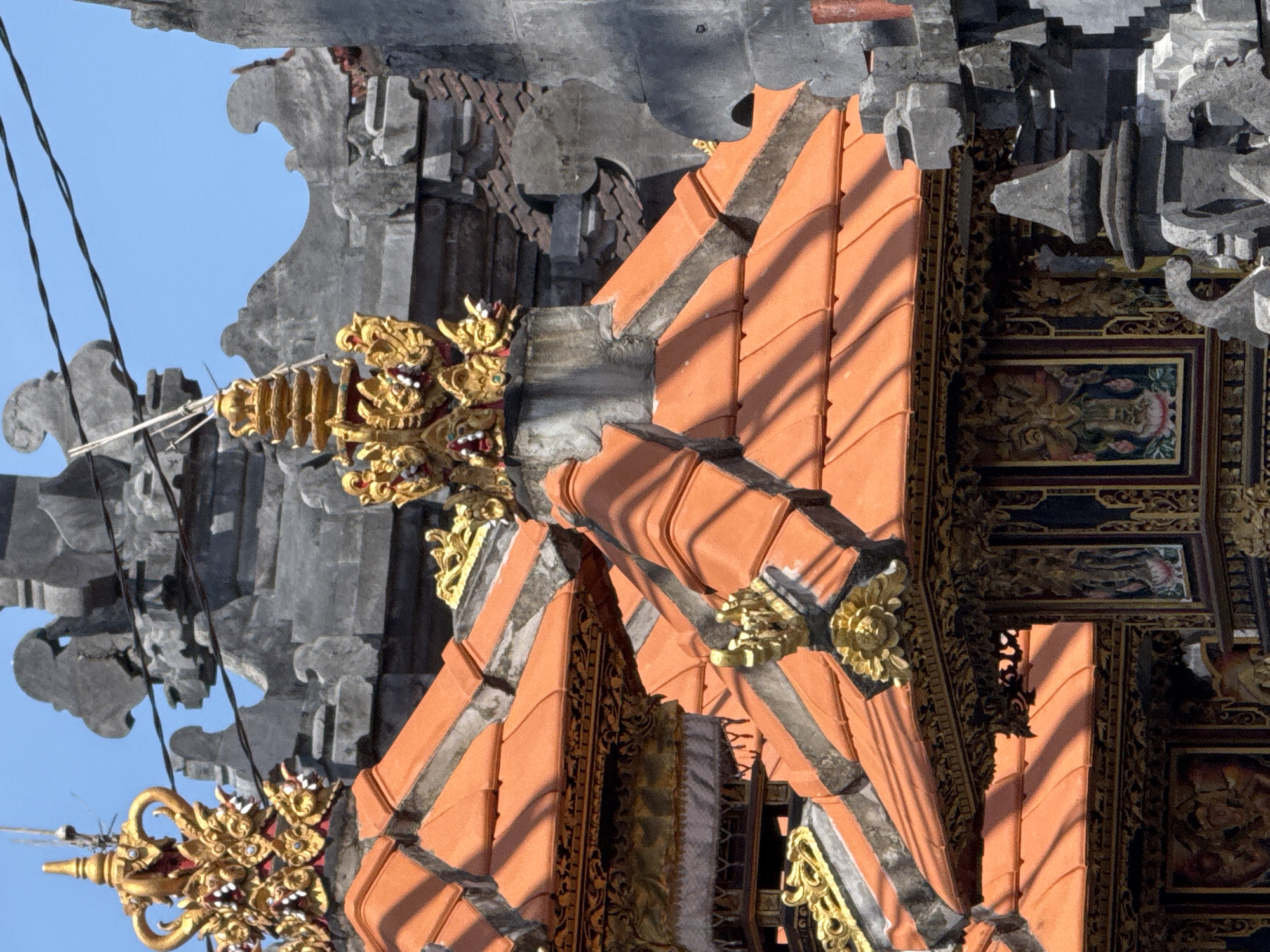 The image shows the exterior of a building in Nusa Dua, Indonesia, featuring a traditional Balinese architectural style. The structure has orange tiled roofs accented with gray trim and intricate gold decorative elements. A detailed gray stone structure with elaborate carvings is visible behind and to the side of the tiled roof. A blue sky is seen in the background, contrasting with the textures and colors of the building's architecture. The scene is well-lit, highlighting the textures and details of the building materials.