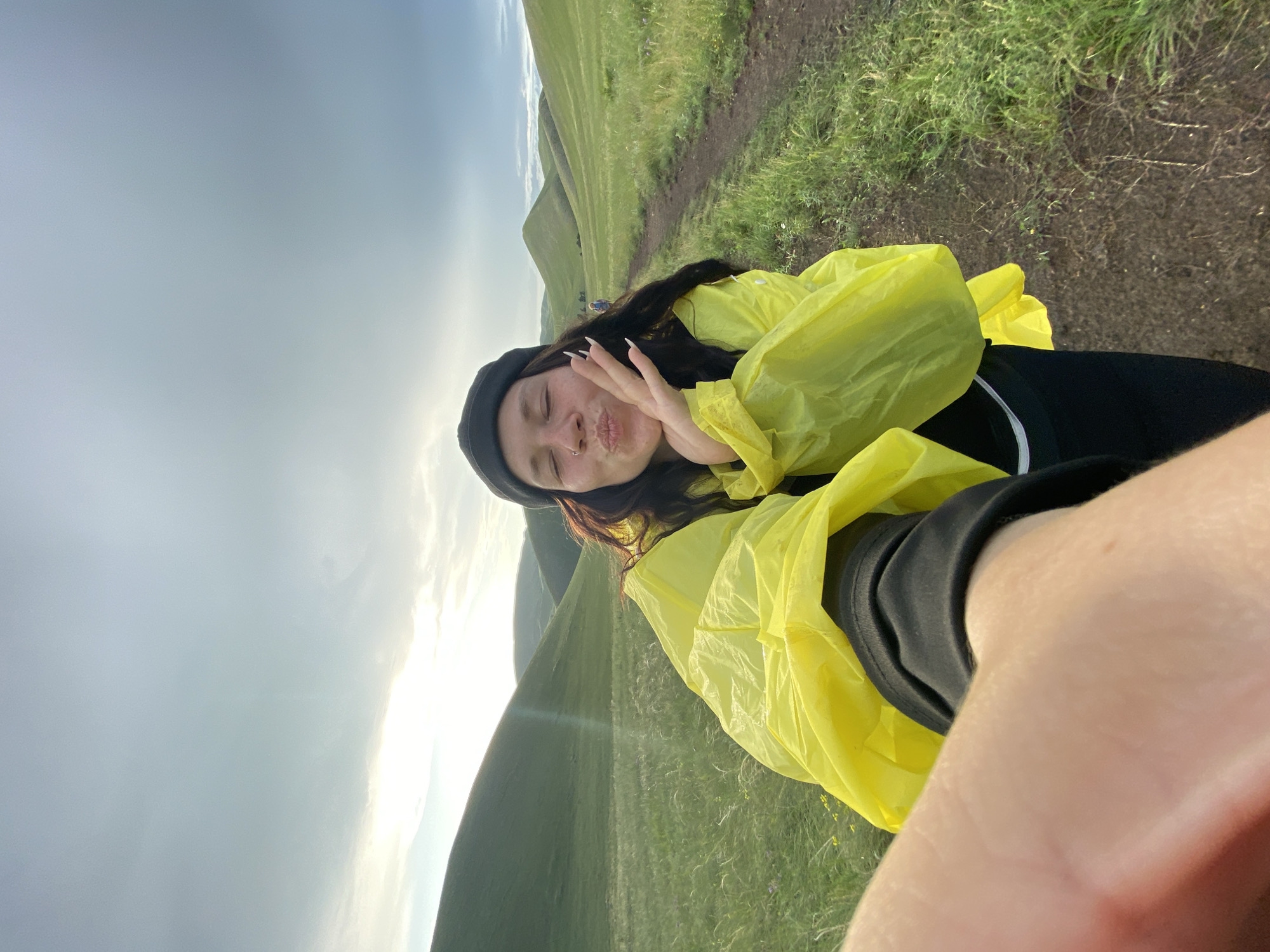 A selfie shows a young woman with long, dark hair wearing a black beanie and a bright yellow rain jacket. She is puckering her lips and holding one hand up to her face, with her eyes closed. She is standing on a dirt path in a grassy, rolling landscape. In the background, hills covered in green grass stretch towards a cloudy sky. The overall impression is of someone enjoying the outdoors, possibly on a hike. The location is the city of Donskoe, Russia.