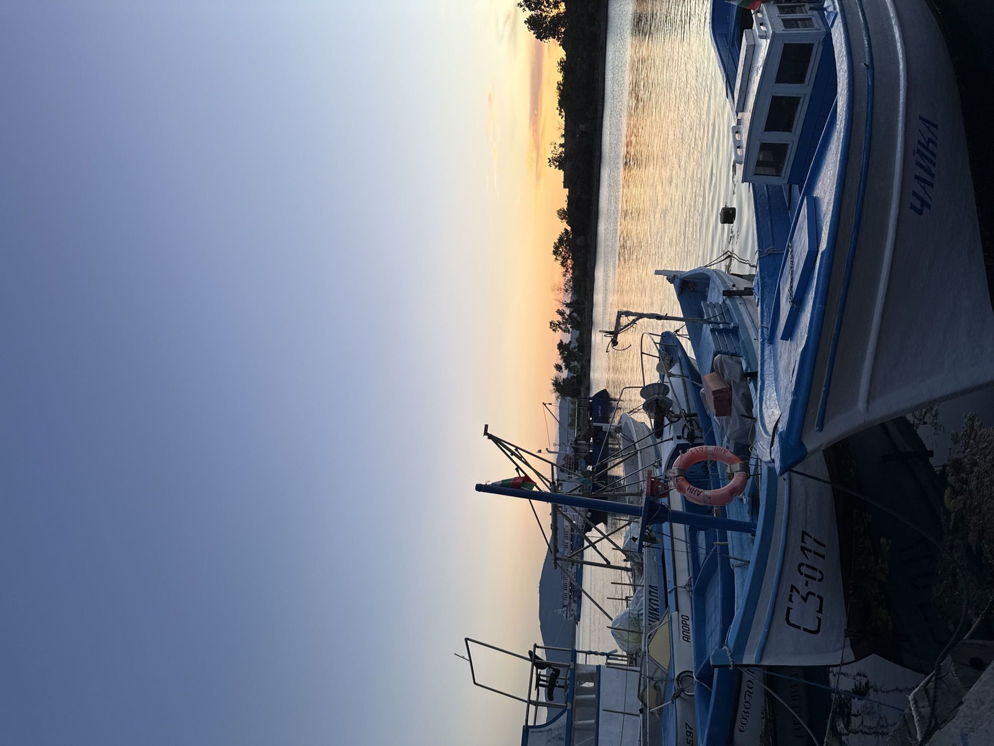 This is a vertical, eye-level photograph of a harbor in Sozopol, Bulgaria, at dusk. The sky is a gradient of clear blue at the top, fading to soft orange and pink hues near the horizon where the sun is setting. A calm body of water, likely the Black Sea, reflects the sky's colors.
In the foreground and midground, several fishing boats are moored together. The boats are predominantly painted white and blue. Some have visible Cyrillic lettering on their hulls. One boat in the center is labeled "C3-017". Another boat in the lower right is labeled "ЧАЙКА". A bright orange life preserver is attached to the railing of one of the boats.
In the background, a tree-lined shore is visible, with silhouetted trees and structures against the colorful sky. A hill with a single structure on its summit is visible in the far left background. The overall scene is peaceful and depicts a typical evening in a Bulgarian fishing town.