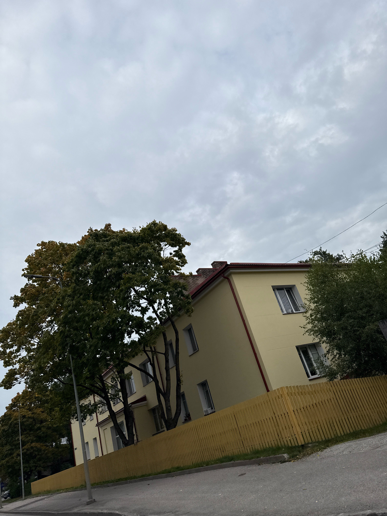 The image shows a building with yellow walls and a red roof in Tallinn, Estonia. The building is a multi-story structure with several windows. In front of the building, there is a yellow wooden fence. Large trees with green and yellow leaves are visible on the left and right sides of the building. The sky is overcast with gray clouds. A paved road and sidewalk are in the foreground.