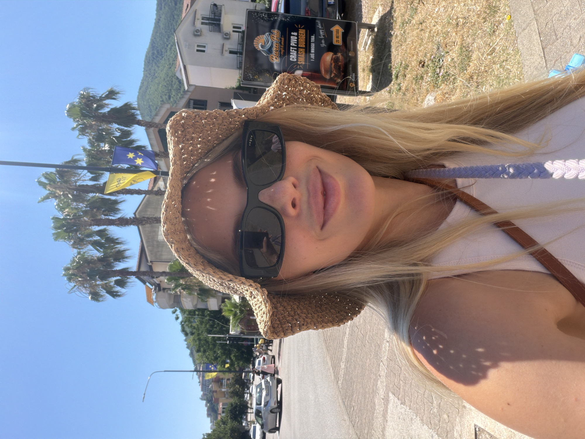 This media file is a vertical selfie of a young woman with blonde hair, wearing a straw hat and sunglasses. She is outdoors on a sunny day, with a clear blue sky and palm trees visible in the background. Behind her, buildings and a street with parked cars can be seen. There is a sign in the background advertising "Bura Rib Craft Pivo & Smash Burgeri". The text on the sign indicates "20 METARA" and "Your happy place". The lighting suggests it is daytime. The woman's facial expression is relaxed and she appears to be enjoying herself. The overall scene suggests a vacation or leisure outing in a warm, sunny location, possibly a coastal town given the palm trees and the style of the buildings.
