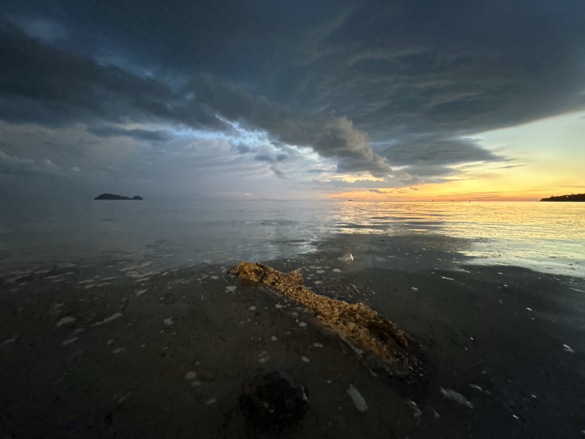 The image depicts a coastal scene at dusk, likely on the island of Koh Phangan, Thailand, given the geotagged location. The sky is dramatic, with dark, heavy clouds dominating the upper portion and a vibrant sunset casting golden and orange hues across the horizon on the right side of the frame. The sea is calm, with reflections of the sky visible on its surface. In the foreground, the wet sand of a beach is visible, with a large piece of driftwood or debris lying partially submerged in shallow water. Small rocks and patches of foam are scattered on the sand. In the distance, a small, dark island or landmass is silhouetted against the sky on the left, and a faint shoreline with vegetation is visible on the right. There are no people or animals clearly visible in the image.