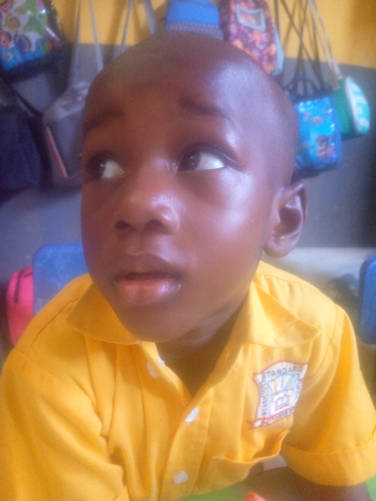 A close-up shot shows a young, bald African boy in a yellow uniform with the "Standard" logo on his chest, looking upwards to the left. The scene appears to be indoors, in a classroom setting in the city of Ikorodu, Nigeria. In the background are several colorful bags hanging. The boy's skin is shiny, suggesting possible perspiration or lighting effects.