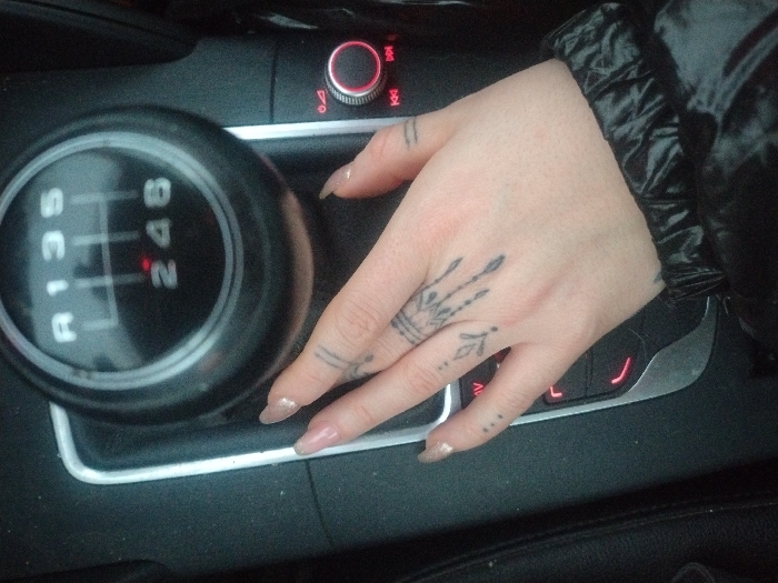 The image provides a close-up view of a car's interior, primarily focusing on a human hand resting near a manual gear stick and part of the center console. The hand, likely female, is adorned with several small, intricate tattoos: a crown-like design on the back of the hand, two parallel lines on one finger, and small dots on another. The fingernails are almond-shaped and coated with a light, sparkly polish. A black, shiny puffer-style jacket sleeve is visible around the wrist.
The prominent black manual gear stick clearly displays the shift pattern for a 6-speed transmission, with "R 1 3 5" and "2 4 6" printed on its top surface. Adjacent to the gear stick, the car's center console features a control knob with a glowing red ring and several other buttons below it, also illuminated in red, set within a silver-trimmed panel. The console surfaces appear somewhat dusty.
The scene depicts a static moment inside a vehicle, with the hand casually placed, suggesting the car is either parked or waiting. The location context provided is Fentange, Luxembourg. No specific time of day or weather conditions are discernible from the image.