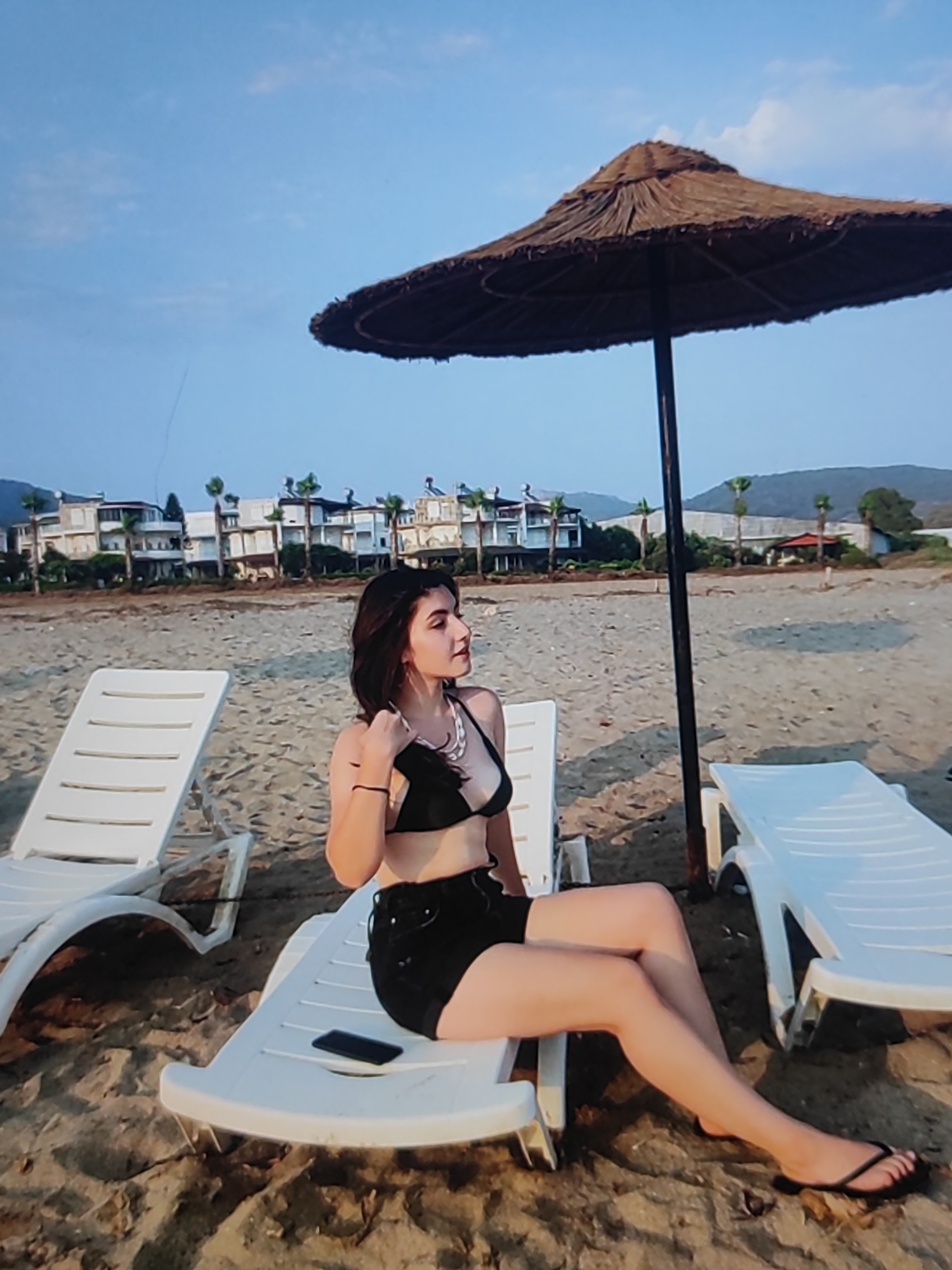 A young woman is relaxing on a beach. She is sitting on a white lounge chair, wearing a black bikini top and black shorts. She is wearing a necklace. Her legs are crossed, and her right foot is resting on the sand. A smartphone is visible on the lounge chair next to her.
Behind her, another white lounge chair is empty. A straw umbrella is set up on the beach, providing shade. The background shows a sandy beach with buildings and palm trees in the distance, suggesting a resort or coastal town. The sky is blue with some clouds, and the lighting suggests it is late afternoon or early evening. The overall mood is relaxed and peaceful.
The location is identified as Fergana City, Uzbekistan. However, based on the visual cues of a beach, lounge chairs, and a resort-like setting with palm trees, this is inconsistent with the typical geography of Fergana City, which is landlocked and located inland. The visual elements strongly suggest a coastal environment. There is no visible text in the image.