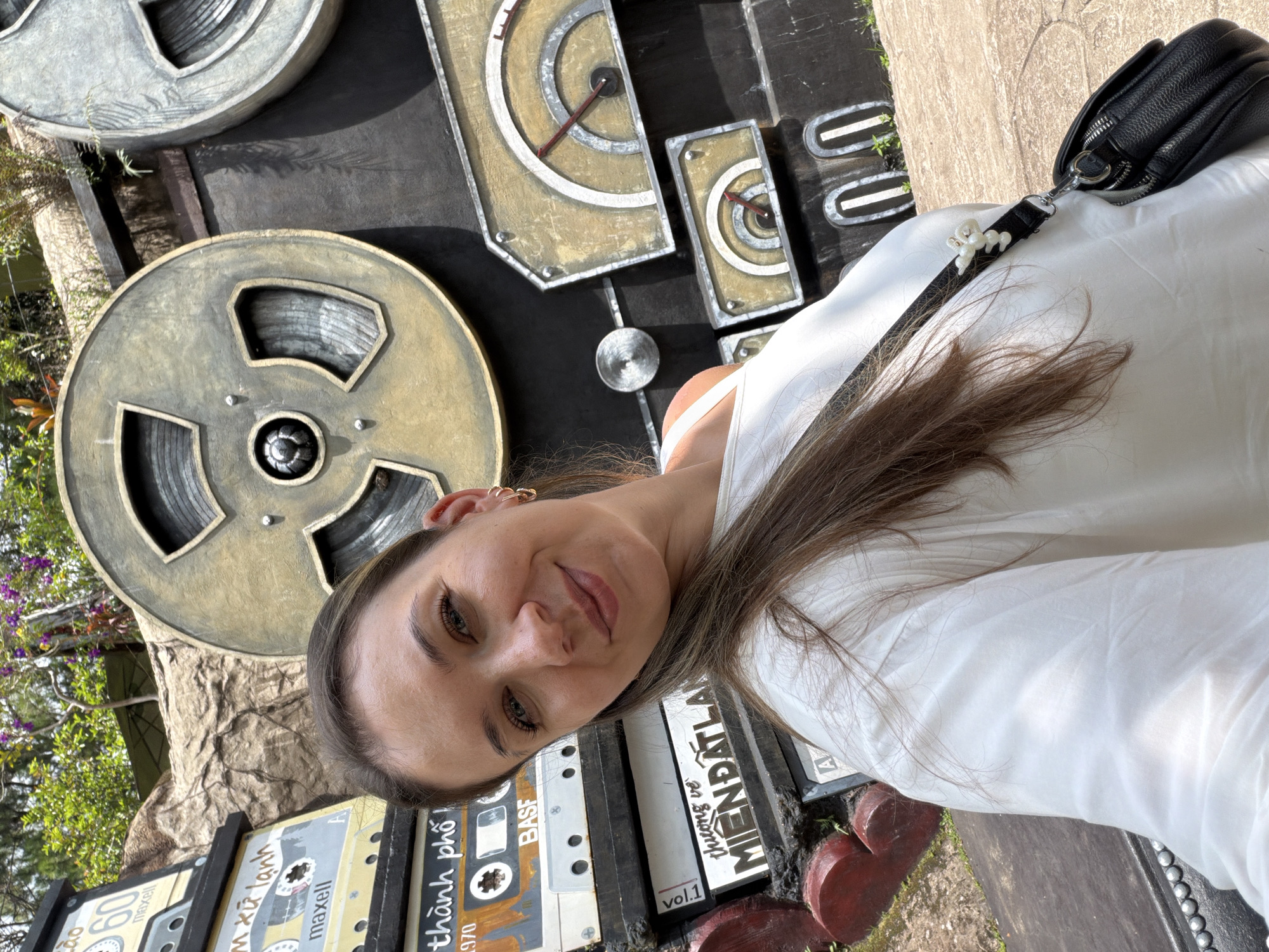 A woman with long brown hair, wearing a white top and a black crossbody bag, smiles at the camera. She is positioned in front of a large, dark wall decorated with what appear to be giant film reels and vintage gauge-like elements. Stacked on the left side of the wall are rectangular objects resembling old cassette tapes and other memorabilia, some displaying text in Vietnamese. The background features lush green foliage and purple flowers, suggesting an outdoor setting. The overall aesthetic of the wall decoration and the presence of Vietnamese text strongly indicate that this location is in Da Lat, Vietnam, likely a thematic installation or art piece.
