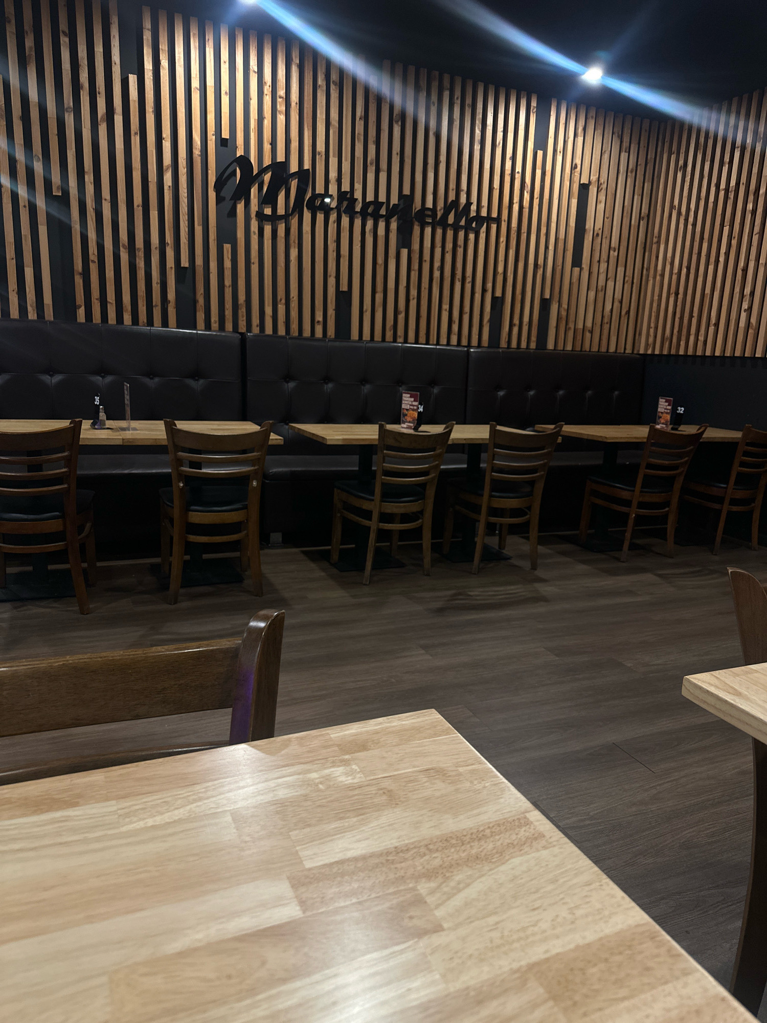 The image depicts the interior of a restaurant, likely in Salisbury, Australia. The dominant visual elements are wooden slatted walls, black leather booth seating, and wooden tables with chairs. A stylized "Moraitis" logo is visible on the slatted wall. Several tables are set with menus and small signs indicating table numbers, such as "32" and "35". The floor is a dark wood laminate. No people are visible in the image. The overall ambiance suggests a modern and clean dining establishment.