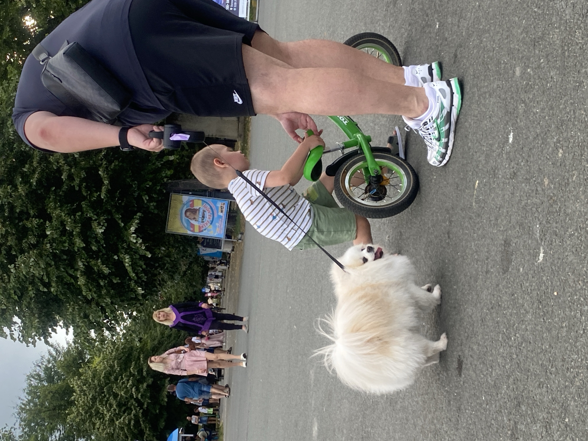 The image depicts an outdoor scene in Burgas, Bulgaria, likely during the daytime. The primary subjects are a man, a young child, and a fluffy white dog. The man is partially visible, wearing black shorts and a dark shirt with a small black bag slung across his shoulder. He is holding onto a small green balance bike, seemingly guiding a young child who is sitting on it. The child is wearing a striped t-shirt and green shorts, actively engaged with the bike. The dog, a small fluffy white breed, is on a leash and walking in front of the child.
In the background, other people can be seen, suggesting a public gathering or park. Two women are visible in the middle ground, one wearing a purple outfit and the other a pink dress. Further back, more individuals are walking and interacting, hinting at a lively atmosphere. Trees and foliage line the background, indicating a green, possibly park-like setting. The ground appears to be asphalt or pavement. A poster with an image of a child and some text in Cyrillic is visible behind the child on the balance bike.
The overall mood appears casual and relaxed, with the focus on the child's enjoyment of the bike and the dog's presence. There are no overt indicators of specific time of day beyond daylight, nor is the weather clearly discernible, though the presence of trees suggests it's not a barren landscape. The scene is likely a public space where families and individuals are spending time outdoors.