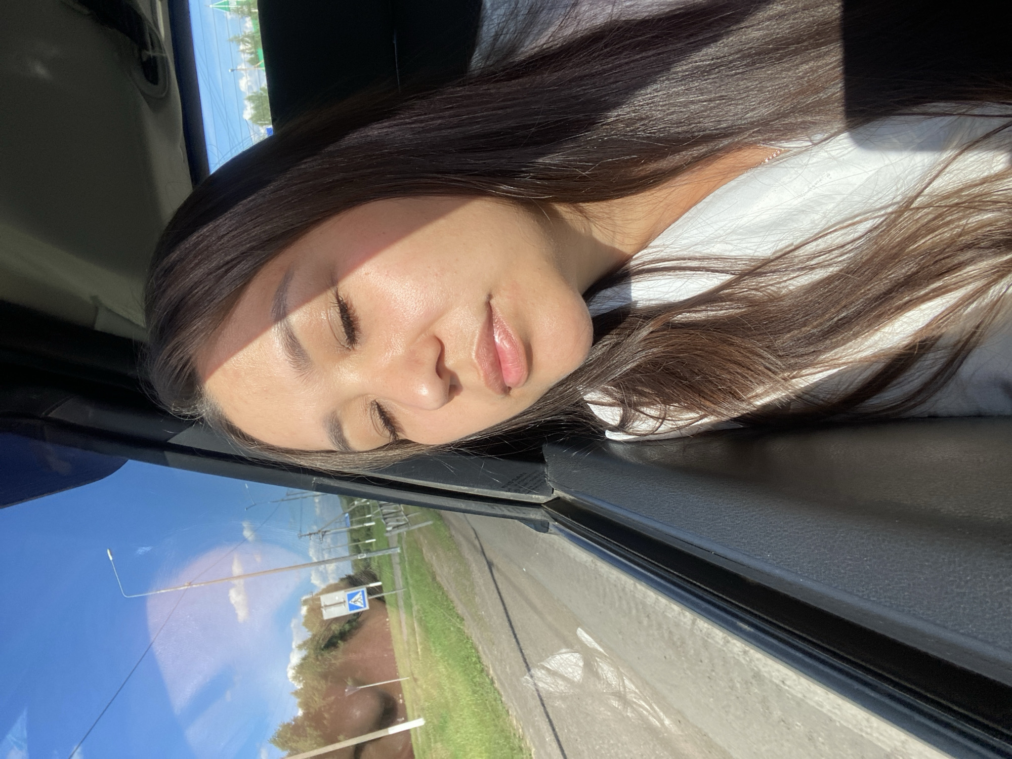 A young woman with long, dark hair is seated inside a vehicle, her eyes closed and her head tilted slightly downwards. Sunlight streams through the window, casting a shadow across her face and highlighting her features. She is wearing a white top. The vehicle's interior is visible, including the door panel and a portion of the window frame. Outside the window, a rural landscape can be seen, with a dirt road, green grass, trees, utility poles, and a blue pedestrian crossing sign under a bright, sunny sky with scattered clouds. The image appears to have been taken in Isyangulovo, Russia, suggesting a journey through a rural area.
