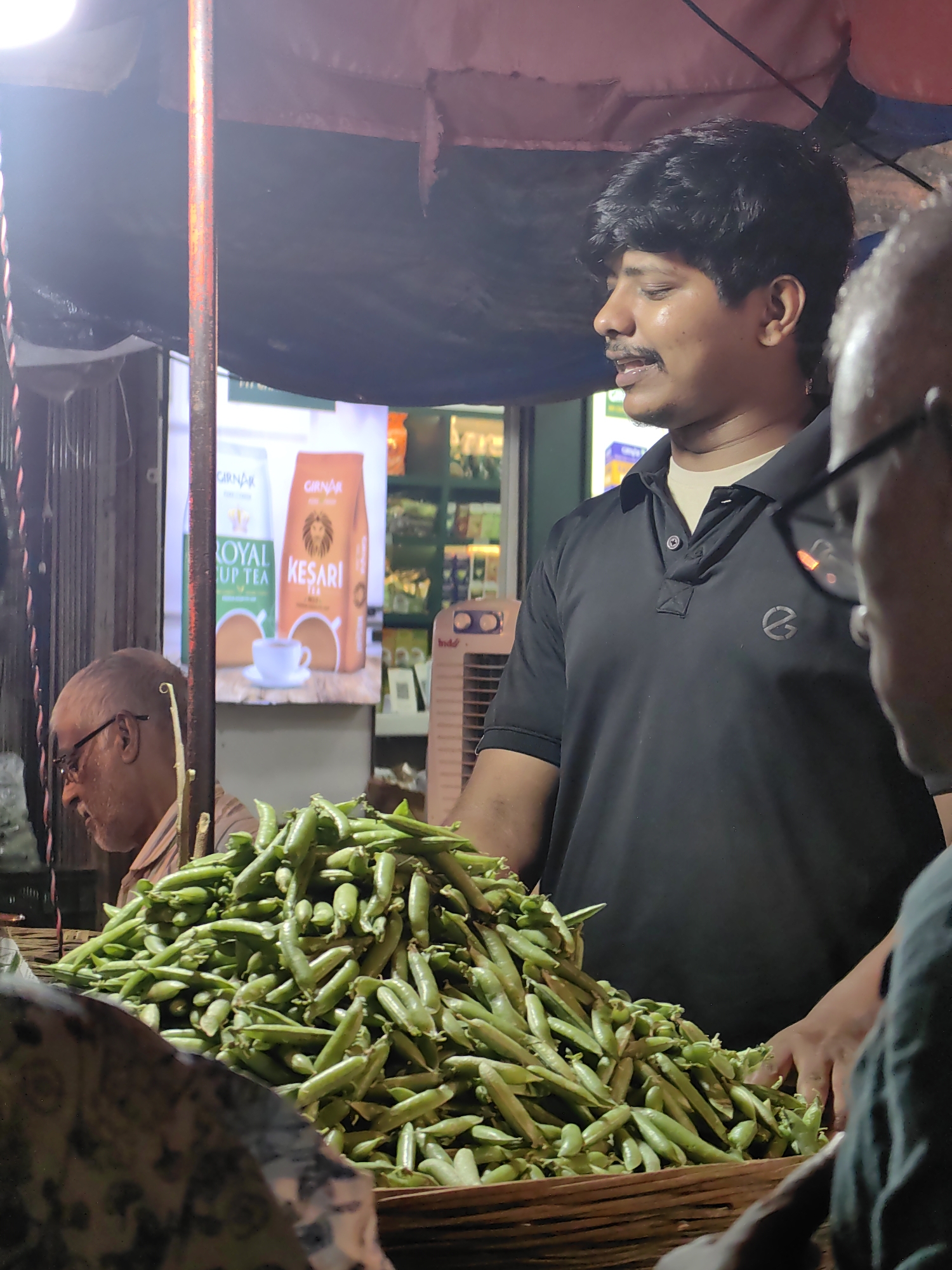 A street vendor, a man with dark hair and a mustache, stands behind a large pile of fresh peas at a market stall in Mumbai, India. He is wearing a black polo shirt with a logo on the left chest and a light-colored undershirt. His right hand is resting on the peas, and he appears to be looking to his left, possibly at a customer. In the background, a poster displays images of tea boxes and cups. To the left of the vendor, an older man with glasses and a striped shirt is also visible. The market stall is lit, with a dark fabric awning overhead and a metal pole supporting it. The overall scene depicts a typical street market environment.