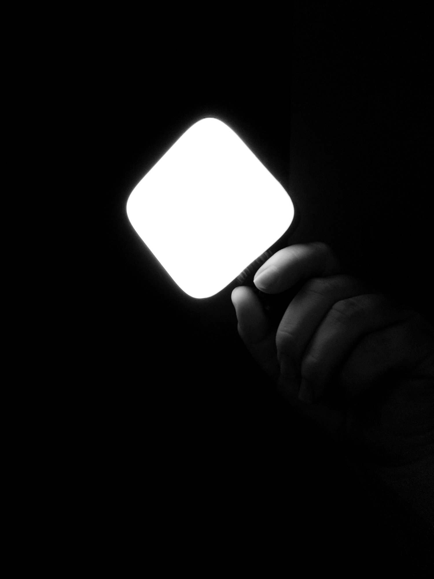 In a dimly lit environment, a hand is holding a square-shaped object that emits a bright light. The object has rounded corners and is positioned in the upper portion of the frame, casting a strong glow. The hand, partially visible and rendered in grayscale, is holding the object from behind and below. The background is predominantly black, emphasizing the luminescence of the object and the details of the hand. The image is a close-up shot, with the focus on the interaction between the hand and the light source. There are no discernible environmental details or contextual clues that would suggest a specific location within Fergana City, Uzbekistan, or any other geographical setting. The scene is stark and minimalist, highlighting the contrast between light and shadow.