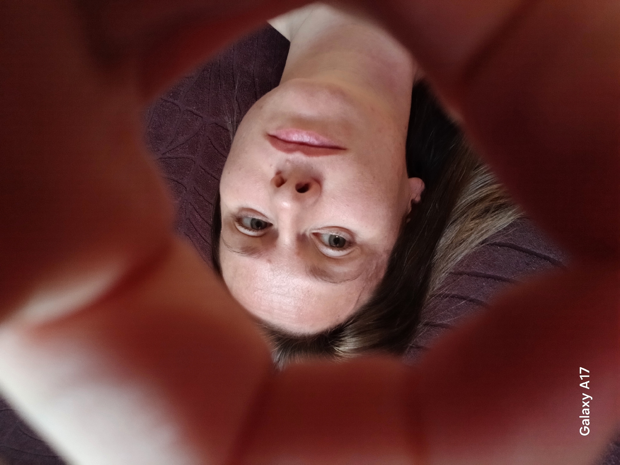 A close-up, over-the-shoulder shot shows a woman looking into the camera, her face framed by blurred red fingers. The woman has brown hair and light skin, with a smattering of freckles across her nose and cheeks. She is wearing light-colored lipstick. The background is a dark brown textured surface. The bottom left corner of the image contains the text "Galaxy A17." The scene appears to be indoors, with no visible indicators of location in Yekaterinburg, Russia.