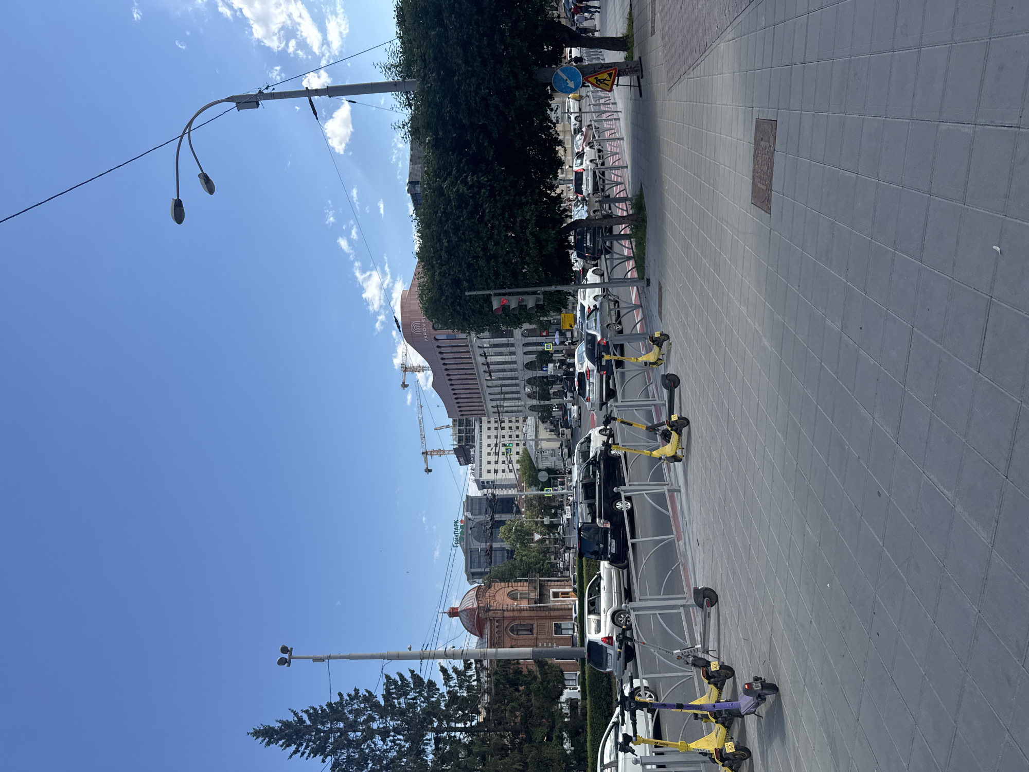 The image depicts a bright, sunny day with a clear blue sky and scattered white clouds. The scene is a city street, possibly in Yekaterinburg, Russia, characterized by a paved sidewalk and a street with parked cars and a row of electric scooters.
On the sidewalk, people are visible walking and standing, suggesting a normal urban day. Several electric scooters, mostly yellow with some purple accents, are parked in a designated area along the street. These scooters are lined up neatly, indicating they are available for rent.
The background features various buildings, including a prominent structure with a domed roof and a large modern building with signage. Construction cranes are also visible in the distance, suggesting ongoing development. Streetlights and traffic signals are present, indicating a functional urban environment. The time of day appears to be midday, given the bright sunlight casting shadows. The overall mood is one of a bustling city on a pleasant day.
There is visible text on some of the buildings and possibly on the scooters, but it is not clearly legible in the provided image. One sign on a building appears to read "Екатеринбург" (Yekaterinburg), and another sign on a scooter might indicate a rental service.