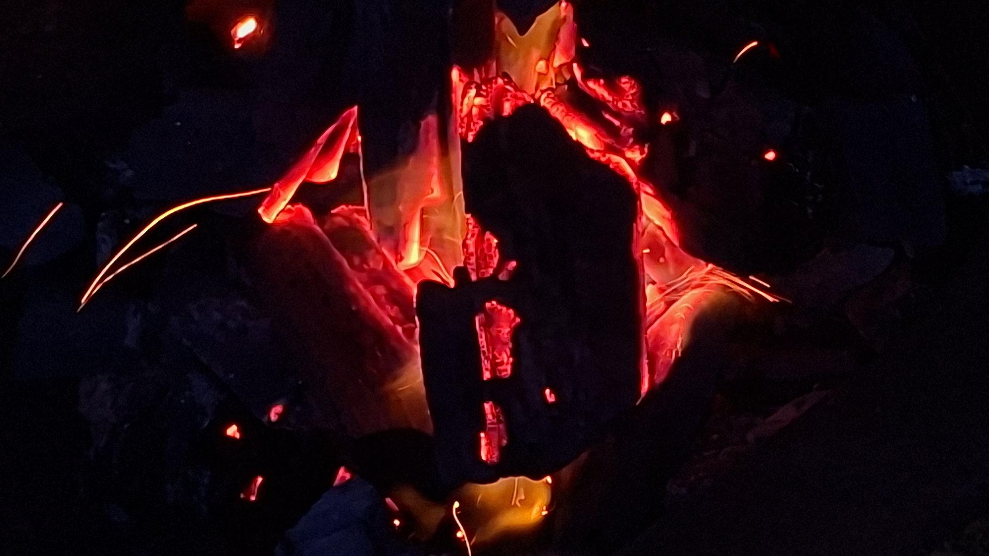 The image shows a close-up of glowing embers and sparks from a fire at night. The scene is predominantly dark, with the intense red and orange hues of the burning material creating a stark contrast against the blackness. The embers appear to be parts of burning wood, with irregular shapes and textures. Fine, bright orange streaks suggest sparks or light trails from the flames. The background is obscured by darkness, but hints of rocky or earthy material can be perceived, suggesting an outdoor setting, potentially a campfire or a bonfire. The overall impression is one of heat and dynamic energy, captured in a moment of intense combustion. No people or specific city landmarks are visible, so it is impossible to determine its exact location within Забалоцкі сельскі Савет, Belarus based on the image alone.