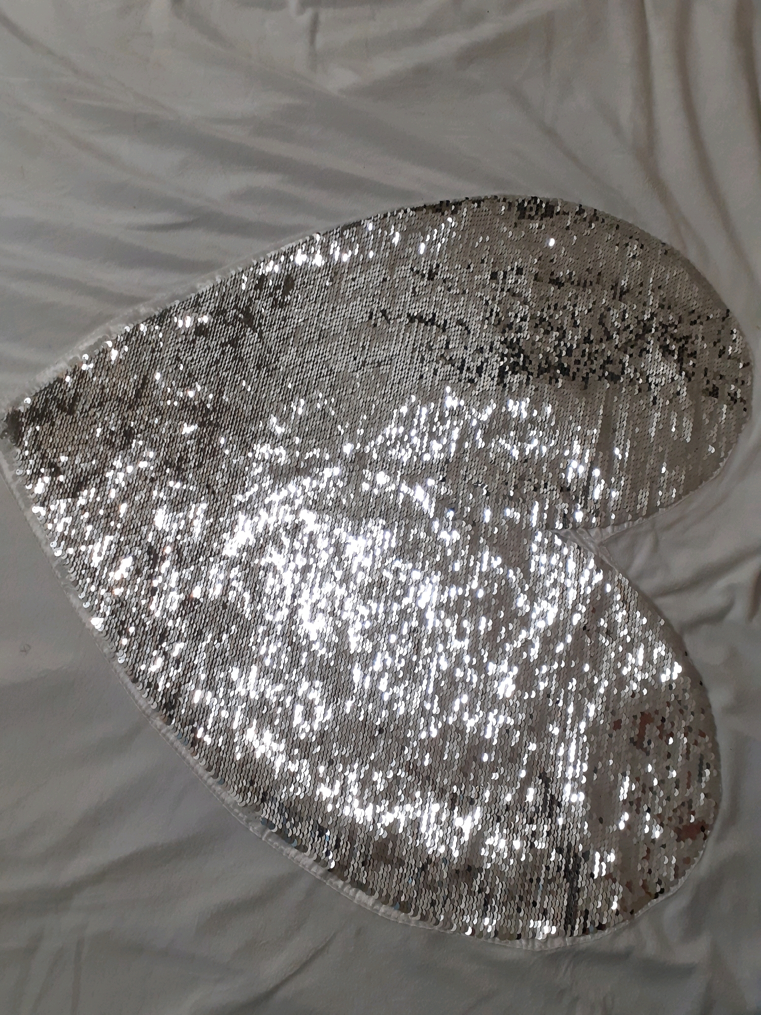 A heart-shaped object with silver sequins is placed on a white, wrinkled surface, possibly a bedsheet or a blanket. The sequins are reflective, creating bright glints of light across the surface of the object. The object appears to be soft and somewhat flat. The background is out of focus and shows folds and creases in the white material. There is no indication of the location being in Tampere, Finland or any specific city.