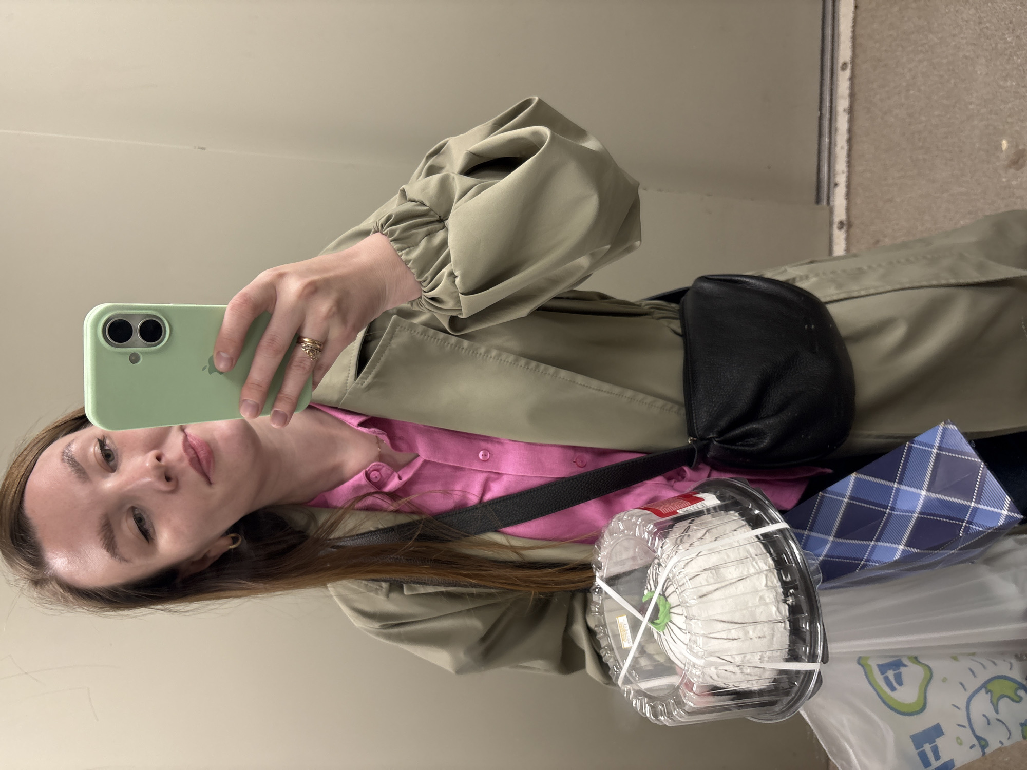 A woman takes a selfie indoors. She is holding a phone with a light green case. She wears a pink button-down shirt under a light green trench coat with puffy sleeves. A black fanny pack is worn across her body. She is carrying a container of what appears to be a cake, and a blue plaid gift bag. A white plastic bag is visible. The setting appears to be a public space, possibly a hallway. There is no visible information indicating the location is Izhevsk, Russia.