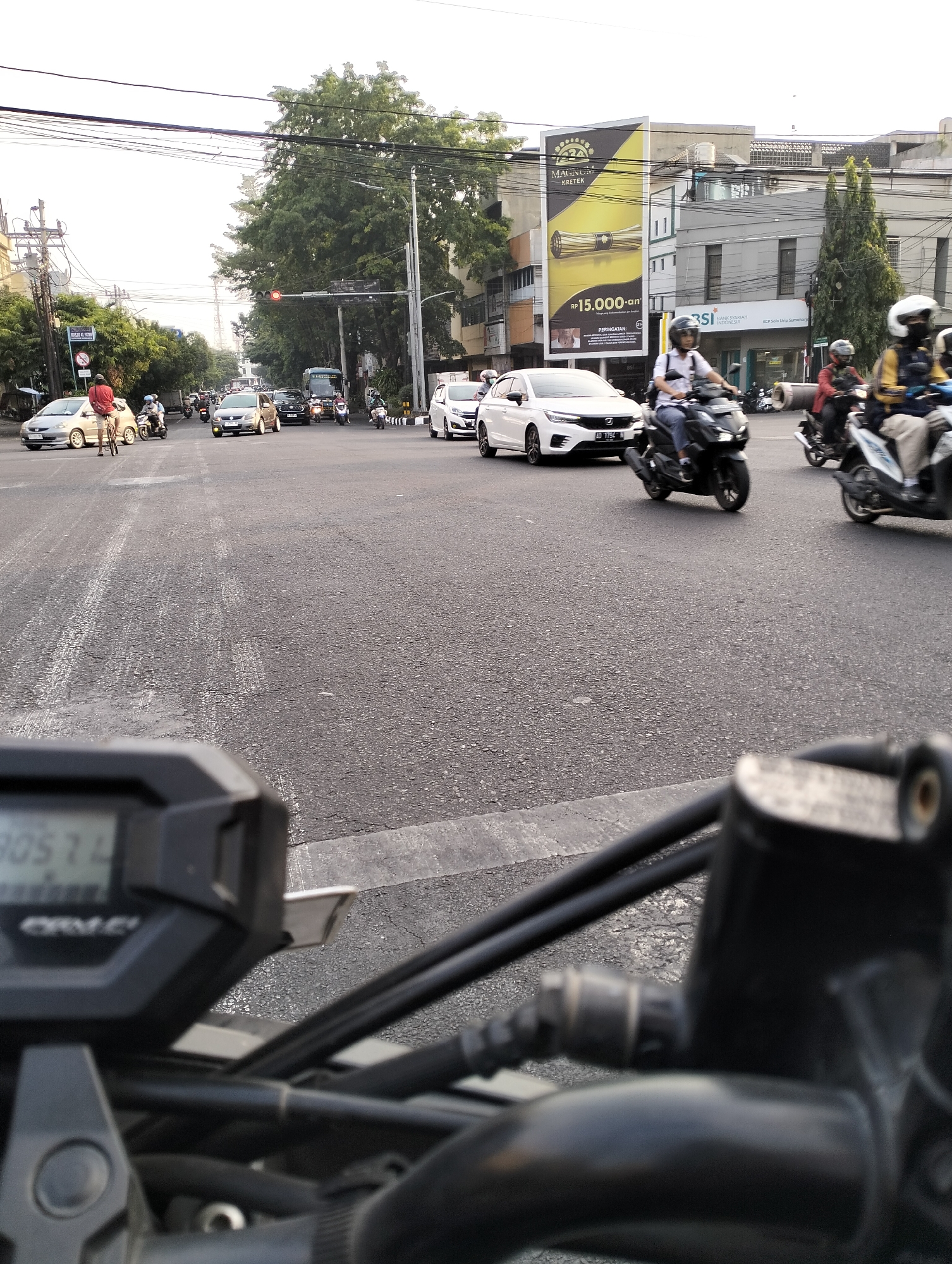 The image captures a street scene in Surakarta, Indonesia, likely at an intersection, characterized by moderate traffic. In the foreground, a portion of a motorcycle's dashboard and handlebars is visible, suggesting the perspective of a rider. The road surface, asphalt, shows lane markings.
Several vehicles are present, including cars and motorcycles. A white car is prominent in the middle ground, followed by other vehicles in motion. A cyclist in a red shirt is also visible, as are multiple motorcyclists, some wearing helmets. Buildings line the sides of the street, with one building displaying a large advertisement with the text "MAGNUM KAIETEK" and a monetary amount in Indonesian Rupiah. Another building has signage for "BSI BANK SYARIAH INDONESIA." Traffic lights are visible, with one appearing to be red. Overhead, power lines crisscross the sky. The overall environment suggests a typical urban setting with active street life and commercial activity. The sky is overcast, and the lighting indicates it is daytime, possibly late afternoon due to the softness of the light.