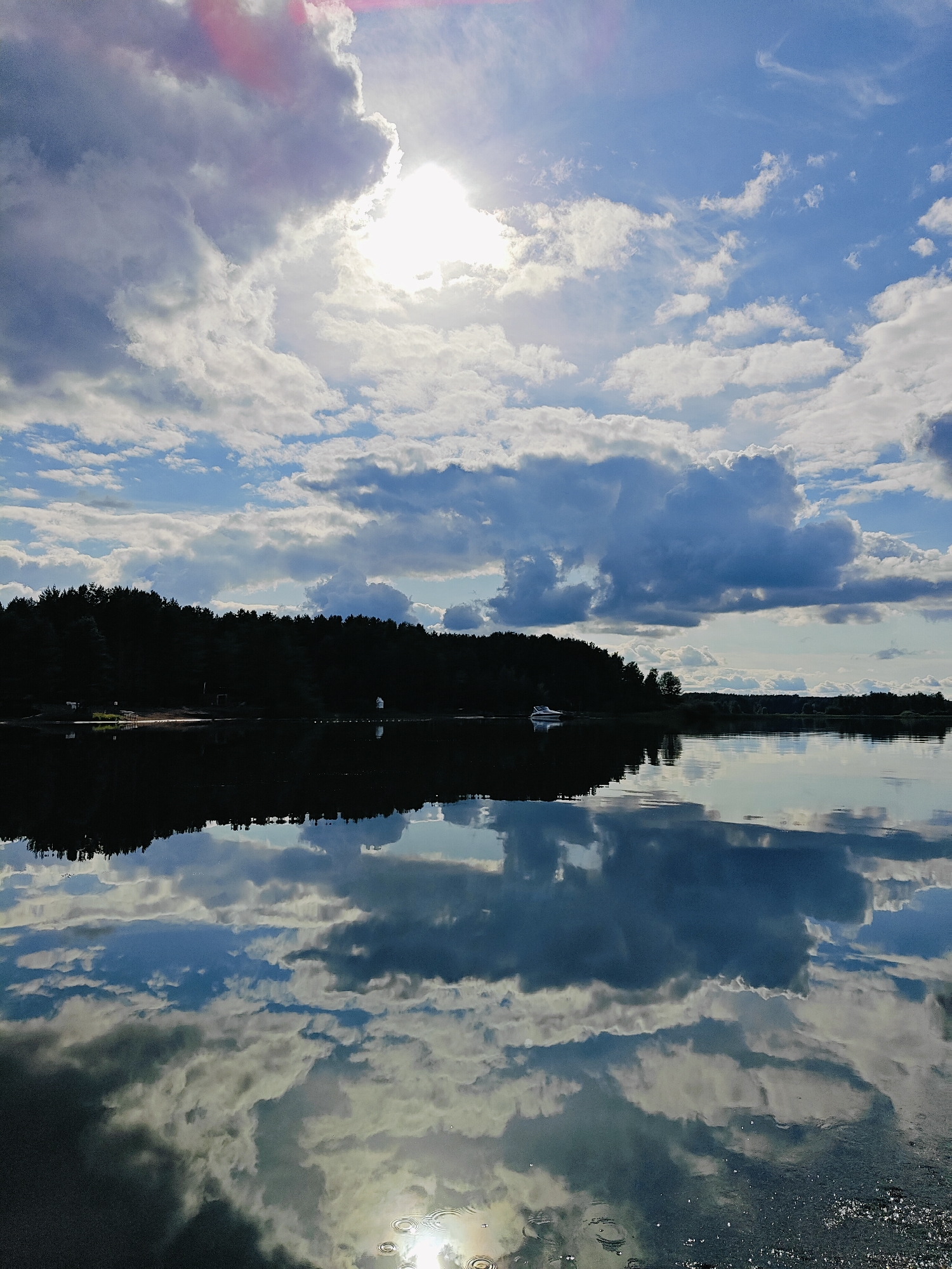 The image depicts a reflective lake with a dark, tree-lined shore under a dramatic sky. The sun is partially obscured by large, white and grey clouds, casting a bright light that creates reflections on the water. A small white boat is visible near the shore in the distance. The water's surface is calm, mirroring the sky and the dense forest along the bank. The overall scene suggests a serene natural landscape. This image does not contain enough information to determine its specific location within the city of Chaykovsky, Russia.