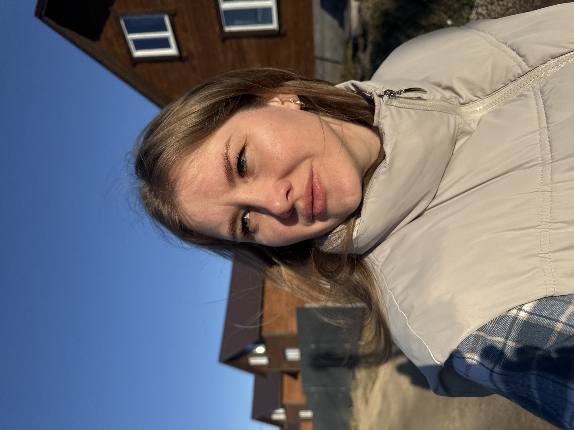 A young woman with fair skin, light brown hair, and blue eyes is the main subject of the image. She is wearing a light-colored puffer jacket over a blue and white plaid shirt. She is looking at the camera with a slight smirk.
Behind her are buildings with brown siding and white-framed windows, under a clear blue sky. The location is Горный, Russia.