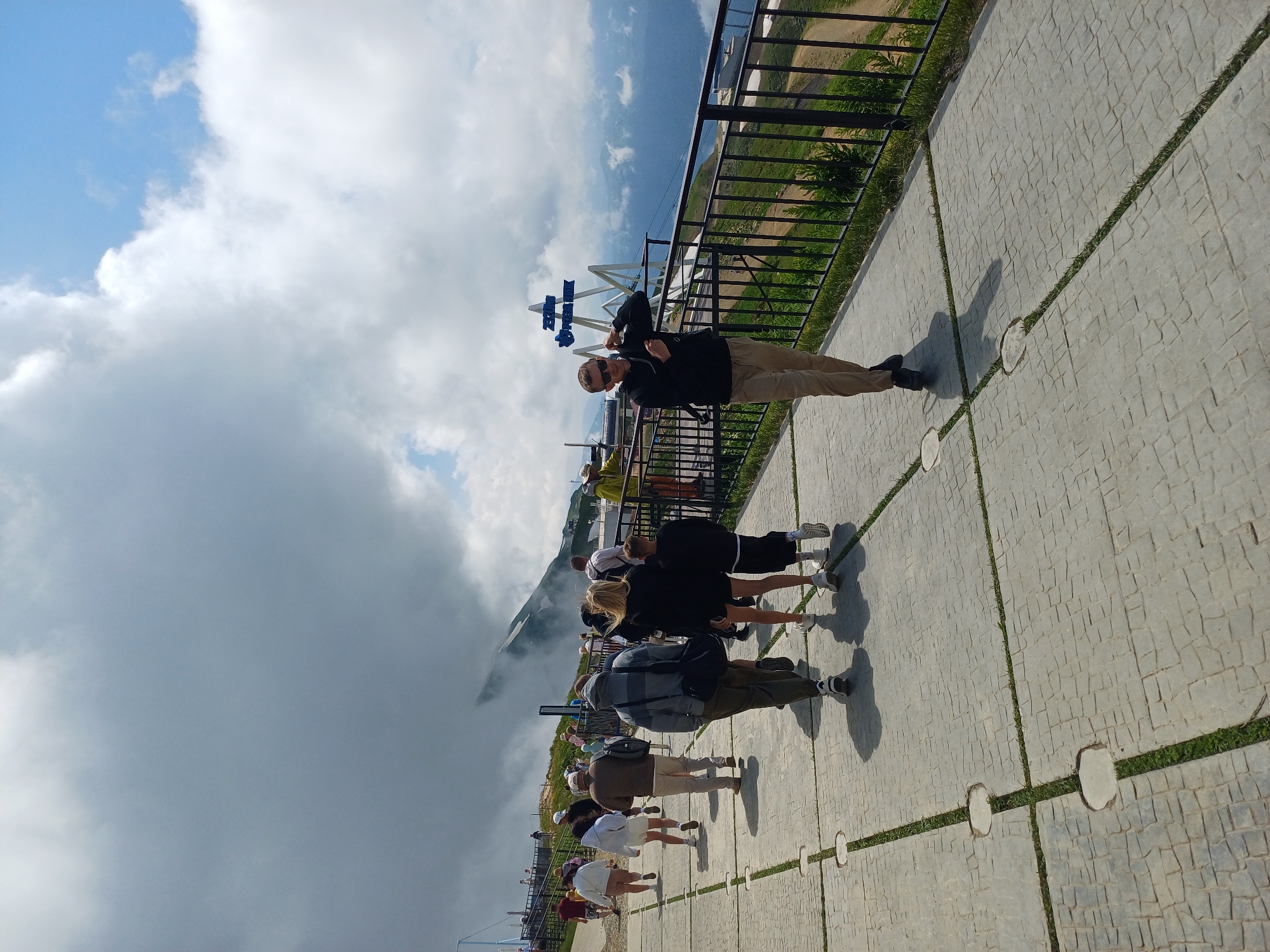 The image depicts a group of people walking on a paved pathway on a mountainside. The pathway is made of large square tiles with grass growing in between the cracks. There is a black metal railing along the right side of the pathway, separating it from a steep drop with a view of mountains and clouds. In the background, there is a sign with blue letters that reads "2320M" and "Rosa Khutor." The sky is partly cloudy with some areas of bright blue and large, grey clouds obscuring parts of the mountains. The overall scene suggests an elevated tourist location with a scenic mountain view.