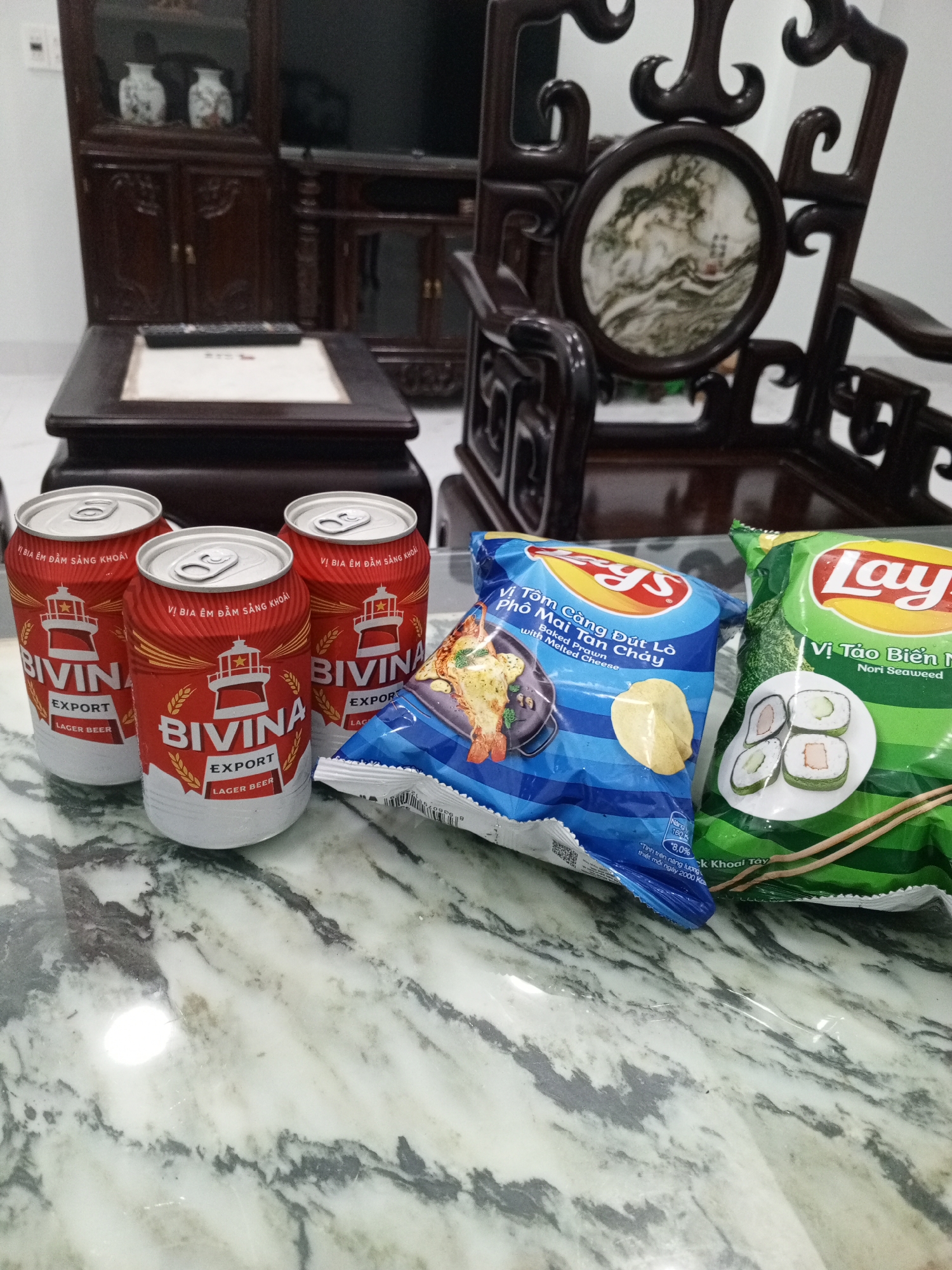 The image shows three cans of "Bivina Export Lager Beer", a bag of "Lay's Baked Prawn with Melted Cheese" flavored potato chips, and a bag of "Lay's Nori Seaweed" flavored potato chips, all sitting on a marble-patterned surface. Behind these items, there's a low, dark wooden table and a dark wooden chair with ornate carvings. A dark wooden cabinet or shelving unit can be seen in the background. The location is Bac Nha Trang Ward, Vietnam.