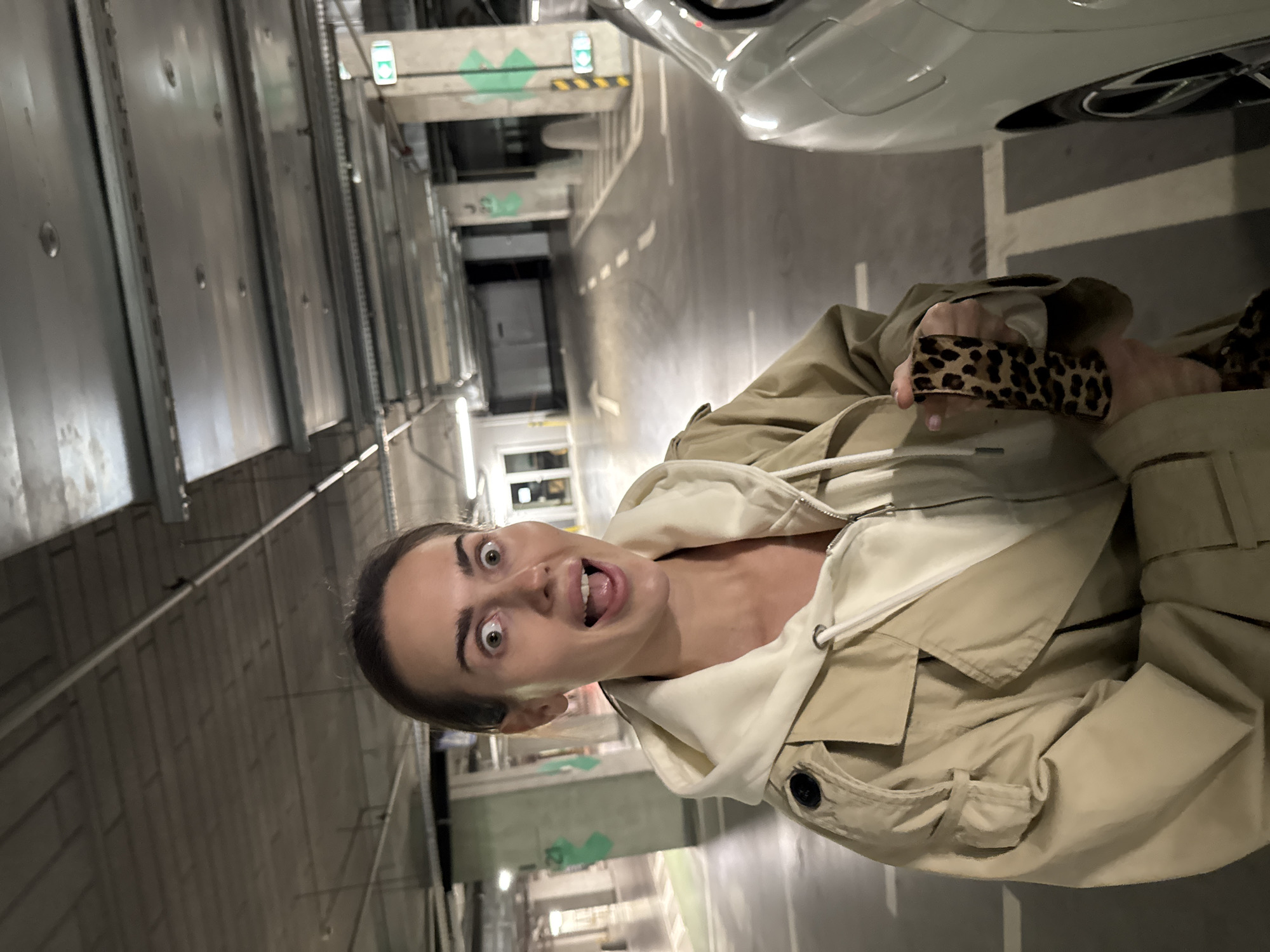 A woman stands in an indoor parking garage, appearing to be near a white car. She is wearing a beige hooded jacket and is holding an object with a leopard print pattern. The lighting in the garage is fluorescent, and the architecture suggests an urban environment. Based on the information available, it's not possible to confirm if the image was taken in Warsaw, Poland.