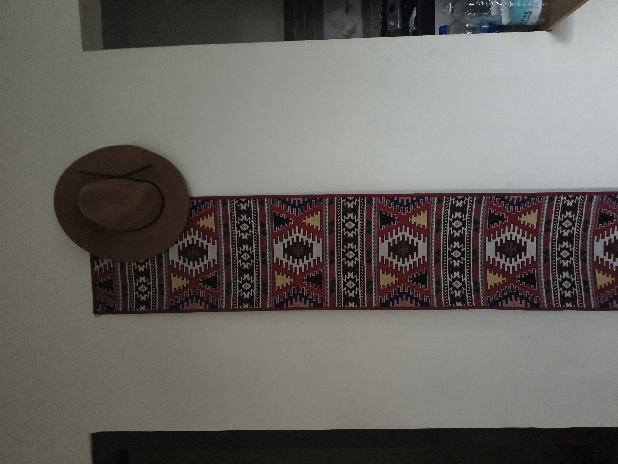 A brown fedora hat hangs on a white wall above a narrow, vertically oriented rug with a geometric pattern. The rug features a repeating design of red, white, black, and yellow diamond shapes. To the right of the rug, a wooden shelf holds several plastic bottles of water. The environment appears to be an indoor setting, possibly a home or a shop. The location is not specified as Haini, India.