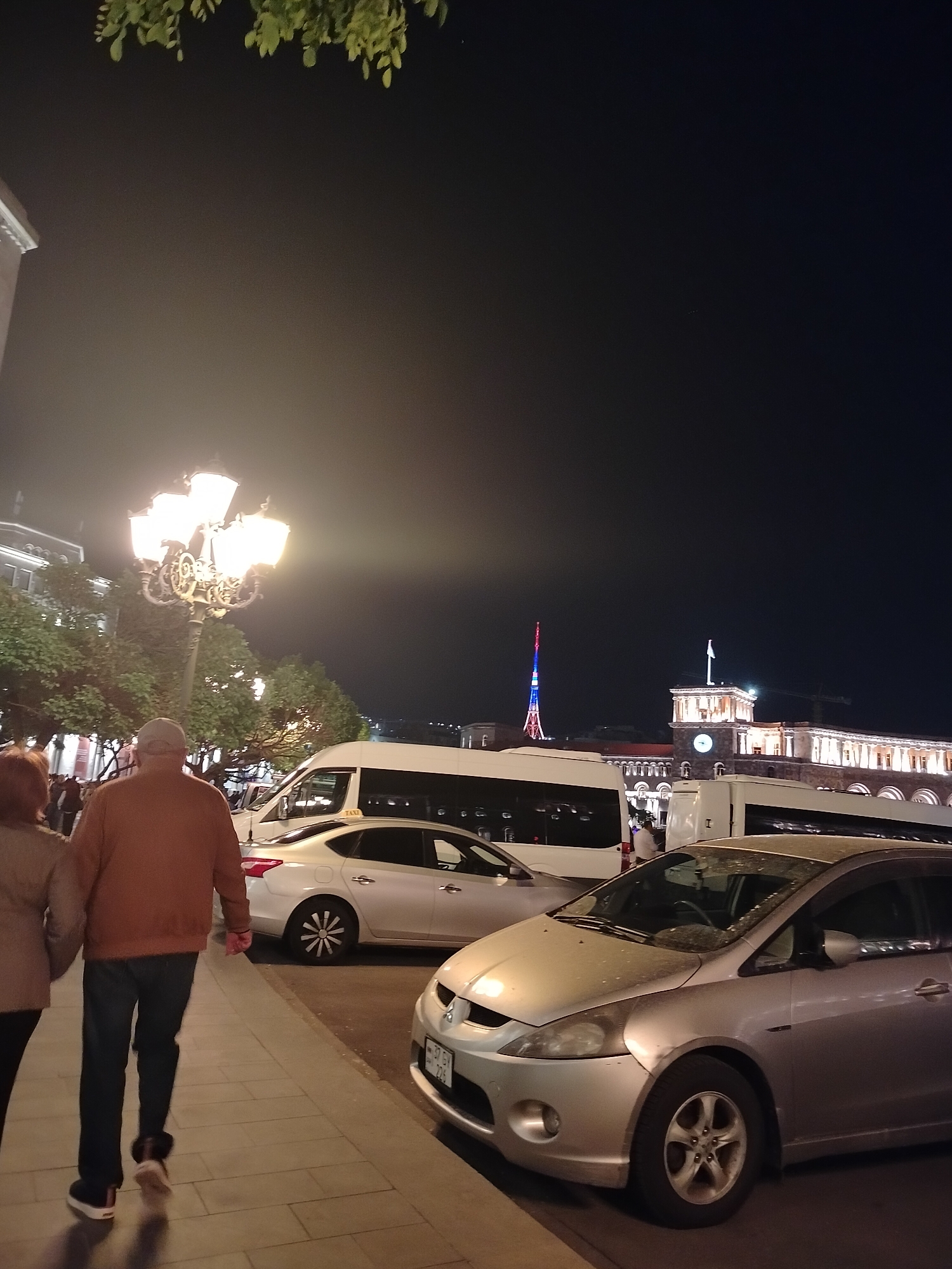 At night in Yerevan, Armenia, an elderly couple walks along a sidewalk lined with parked cars. The man wears a brown jacket and blue jeans, while the woman is partially visible, wearing a light-colored jacket. A tall, ornate lamppost illuminates the scene. Behind the cars, a white van with "TAXI" printed on its side is visible. In the background, the Republic Square building is seen with a flag atop, and a tower is illuminated with red, blue, and orange lights.
