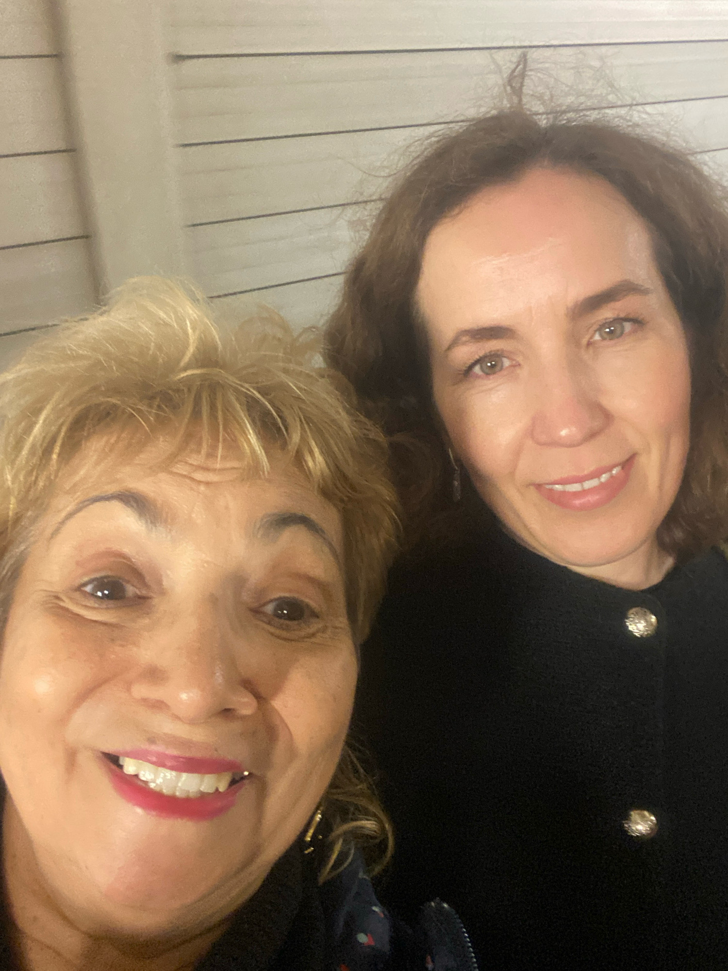 Two women are taking a selfie. The woman in the foreground has short, blonde, curly hair and is smiling with her mouth open. She is wearing a dark top with a blue and red patterned scarf. The woman in the background has long, wavy, brown hair and is also smiling. She is wearing a black top with decorative buttons. The background appears to be a wall with horizontal lines. The lighting is warm and somewhat dim. There is no indication of the location within Moscow, Russia.