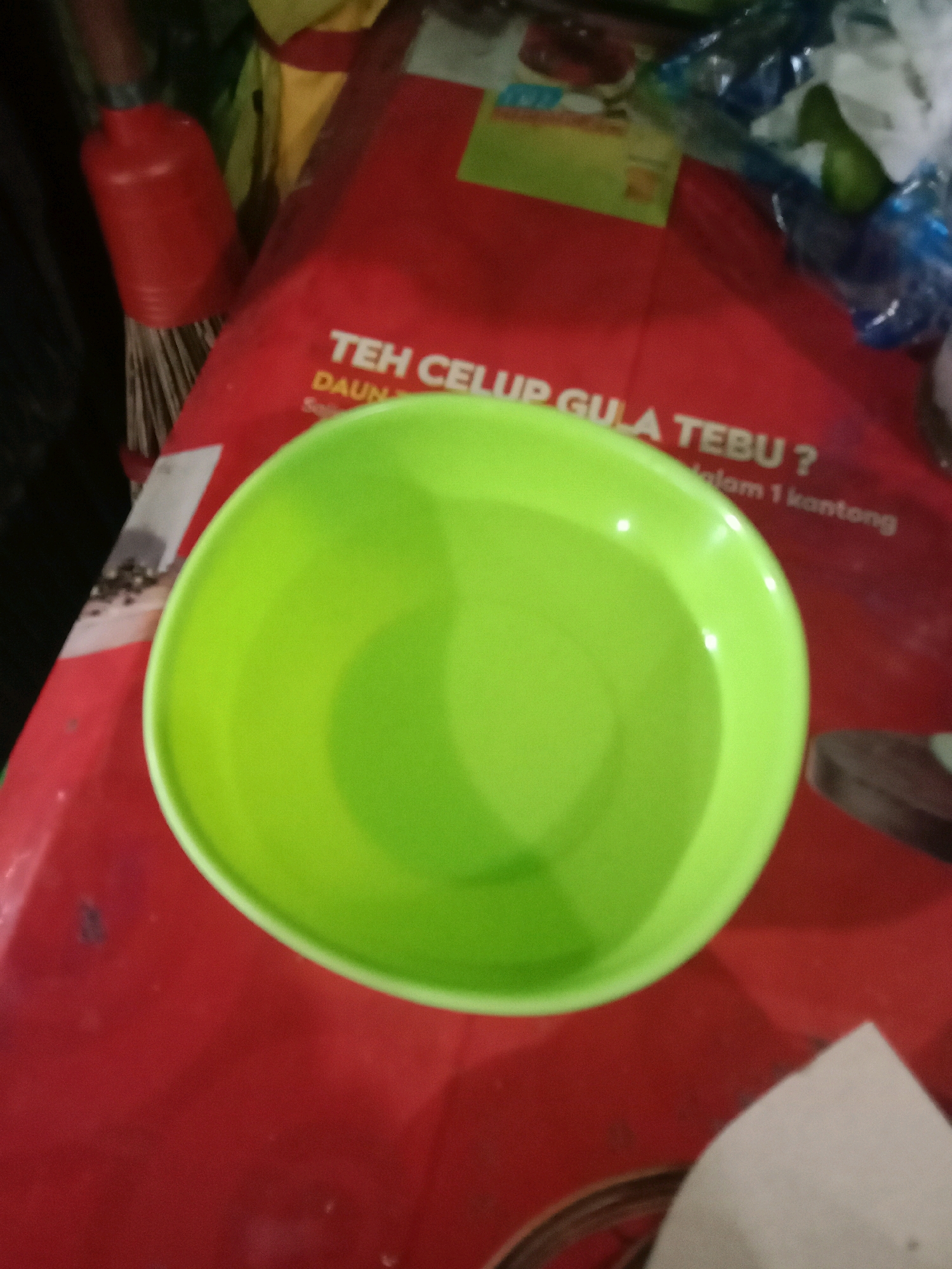 An overhead, slightly blurry shot depicts a bright green plastic bowl filled with clear liquid, positioned on a red surface. The red surface appears to be a table or tray with some text printed on it. The text is partially visible and appears to be in Indonesian, with phrases like "TEH CELUP GULA TEBU?" (Sugar Cane Tea Bags?) and "dalam 1 kantong" (in 1 sachet) discernible. To the left of the bowl, a red object with bristles, possibly a brush or broom, is visible. The background is dark and out of focus, with hints of other objects and packaging. The overall scene suggests a domestic or retail setting, possibly in Semarang, Indonesia, given the language on the packaging.