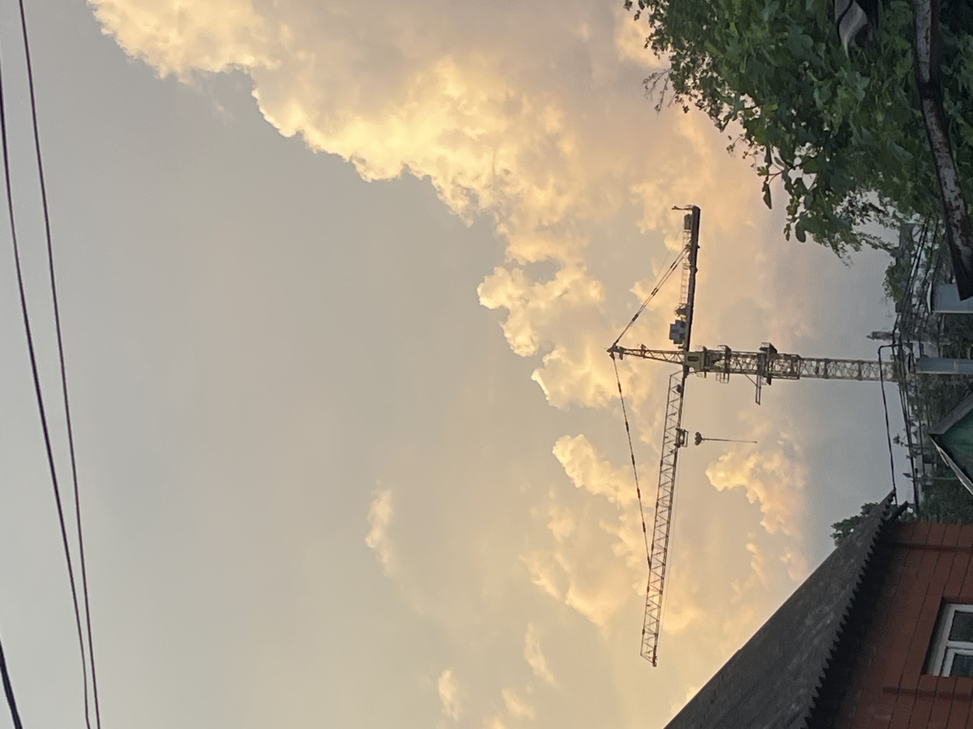 The image captures a construction crane against a sky filled with dramatic, sunset-colored clouds. The crane is a prominent subject, its long horizontal arm extending across the frame. Below the crane, the rooftops of residential buildings are visible, some appearing older with corrugated metal roofs, while others are less distinct. Lush green foliage, likely trees and bushes, frames the right side of the image, suggesting an area with some vegetation within or near a more urban setting. Overhead, thin dark wires are visible, indicating the presence of power lines. The overall scene suggests a transitional period, likely dusk, with the warm glow of the setting sun illuminating the clouds. The image appears to be taken from a low vantage point, looking upwards towards the sky and the construction site. It is difficult to ascertain the exact location in Krasnodar, Russia, without further context.