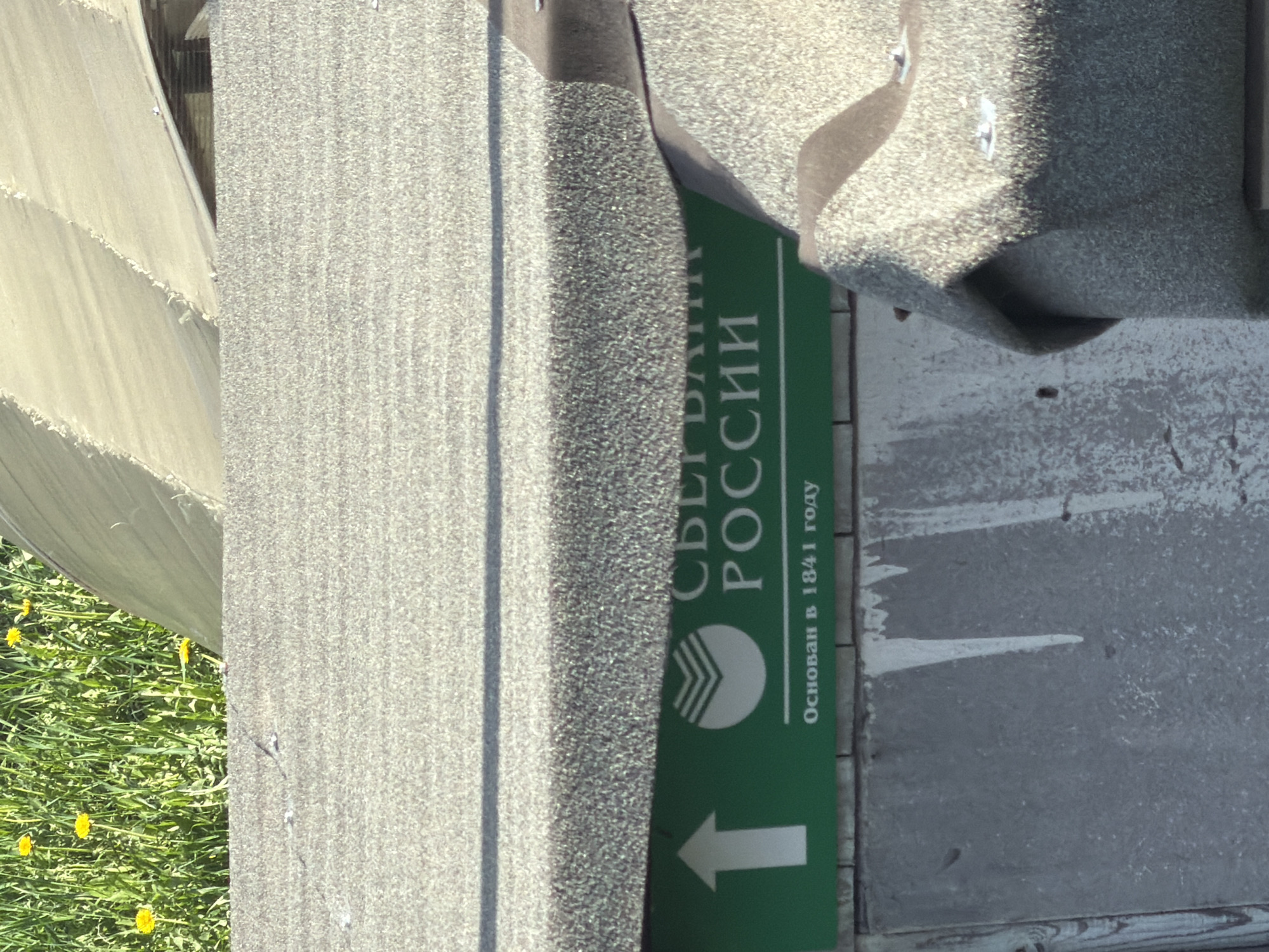 A close-up, sideways view captures a street sign for Sberbank Russia, indicating its location in Plamya, Russia. The green sign features the Sberbank logo, an arrow, and text stating that the bank was established in 1841. The sign is affixed to a weathered, grey concrete structure. Adjacent to the concrete is a rough, possibly abrasive, grey material. A glimpse of green grass and yellow flowers suggests an outdoor setting. The image is brightly lit.