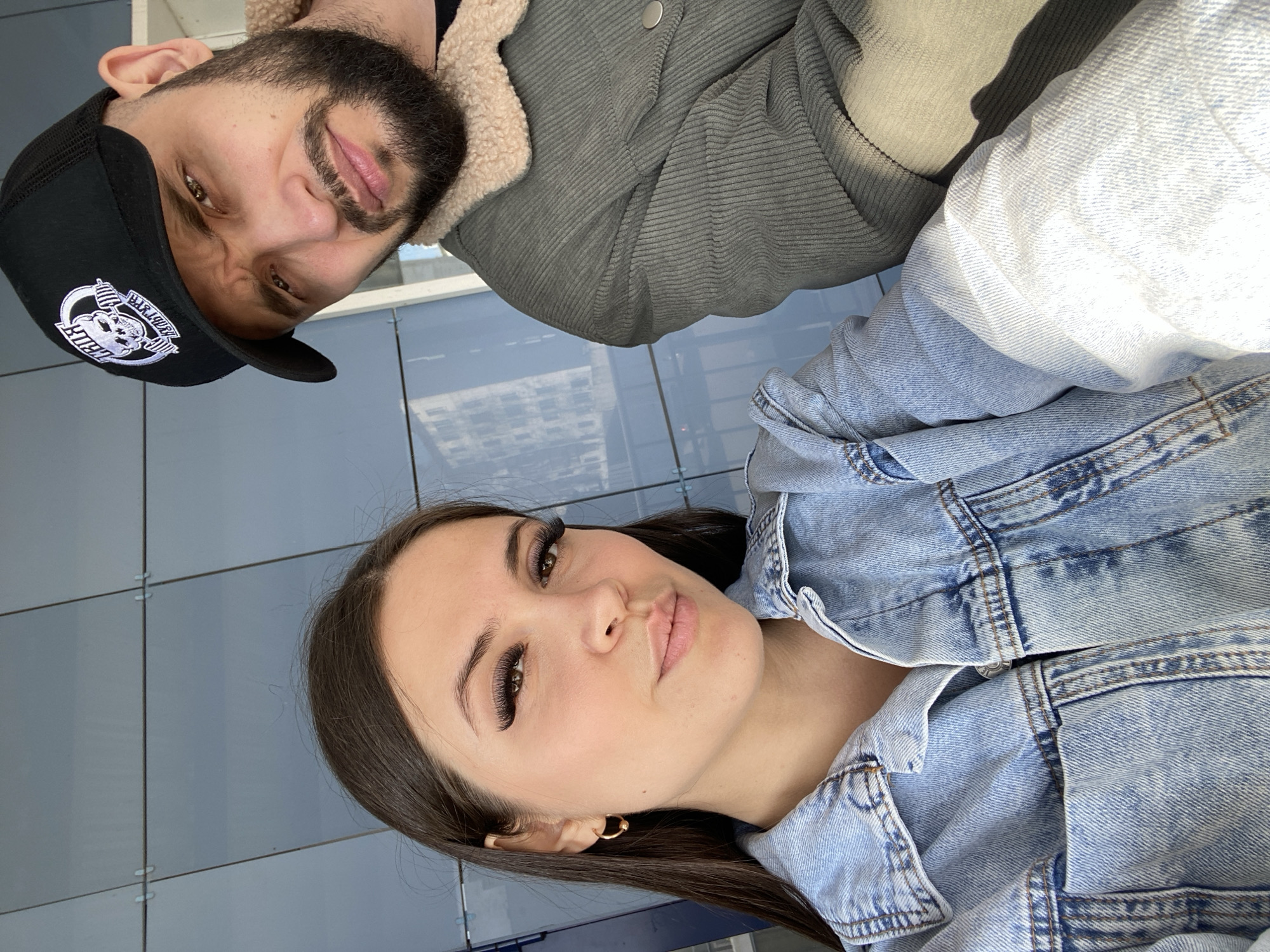 This media file captures a close-up, rotated selfie featuring two individuals, a man and a woman, in what appears to be an urban outdoor setting in Chaikovsky, Russia. The image is rotated clockwise, so the subjects appear sideways.
The man, positioned on the left side (when viewed upright), has a dark beard and mustache, and dark hair. He is wearing a black baseball cap with a white logo that includes the word "Barbell" and an emblem of a figure lifting weights. He is dressed in a dark greenish-grey corduroy jacket with a cream-colored sherpa lining. His expression is a slight, playful grimace or smirk, as he looks directly at the camera.
The woman, on the right side of the frame (when viewed upright), has long, dark hair and prominent eyelashes, possibly extensions. She is wearing a light blue denim jacket and a small golden hoop earring is visible in her right ear. Her lips are slightly pursed in a "duck face" or kissy face pose, and she also looks directly at the camera.
The background consists of blue-grey, rectangular panels or tiles, which could be part of a modern building facade or wall. There are subtle reflections of distant buildings visible on this surface, indicating an urban environment. The lighting is bright and natural, suggesting it is daytime with clear, sunny weather. Both individuals appear to be engaged in a casual, friendly selfie or close-up photo opportunity.