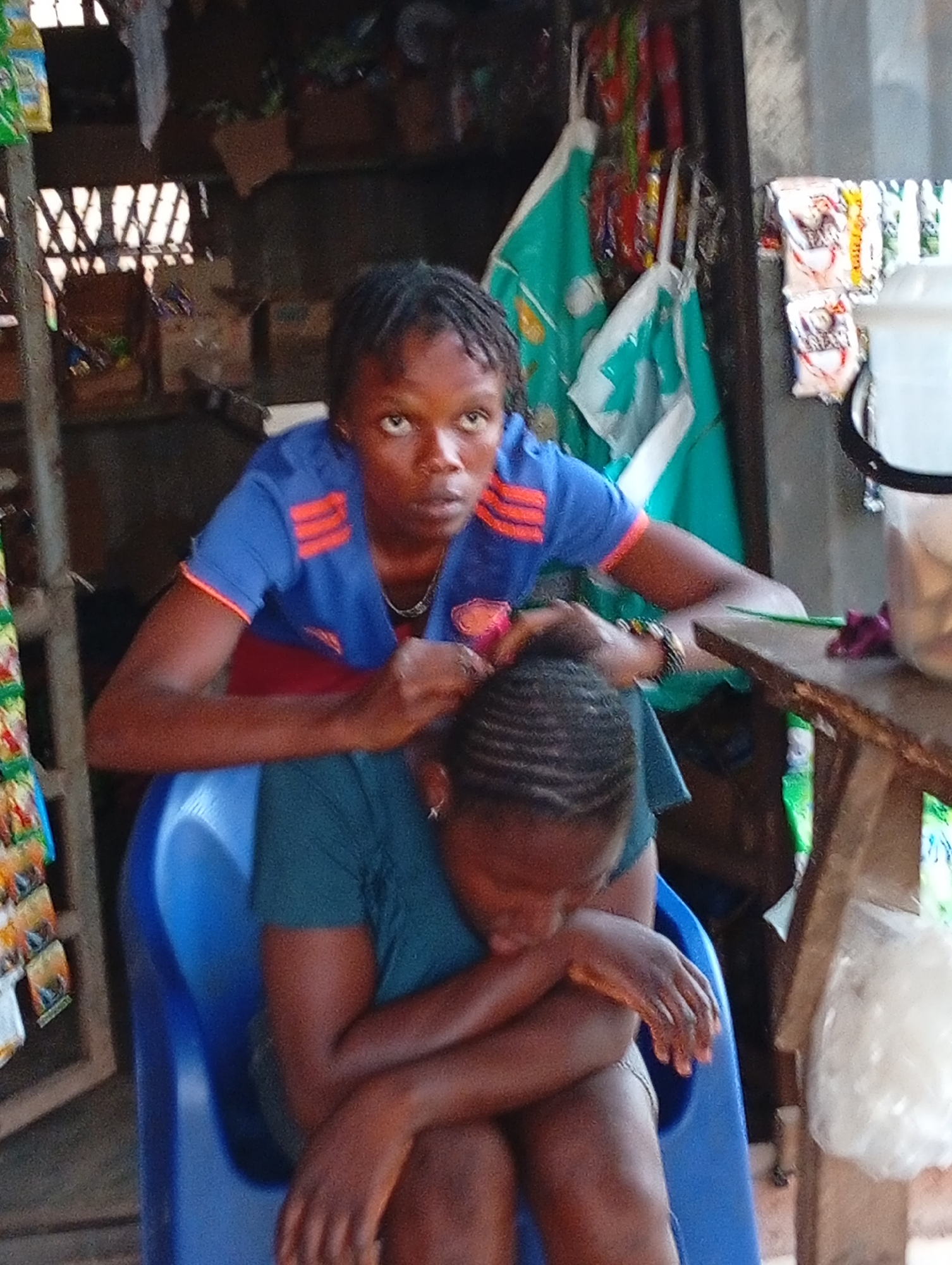In Ogidi, Nigeria, a woman in a blue shirt with orange accents stands behind a seated woman in a green shirt, braiding her hair. The seated woman has her arms crossed and her head bowed. They are positioned in front of a makeshift storefront or stall. Shelves display various items, including packaged goods. A table is to the right, and green cloths hang in the background. The braiding action is in progress.