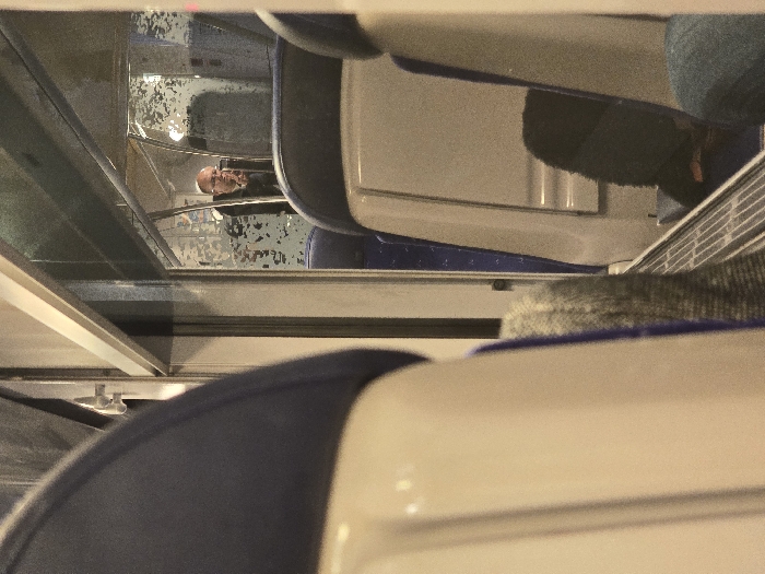 A partial view inside a train car shows rows of seats, predominantly white and blue. Reflections in a glass partition reveal a bald man with glasses wearing a dark shirt. Overhead luggage racks are visible. The seating configuration suggests a public transportation setting, likely a passenger train.