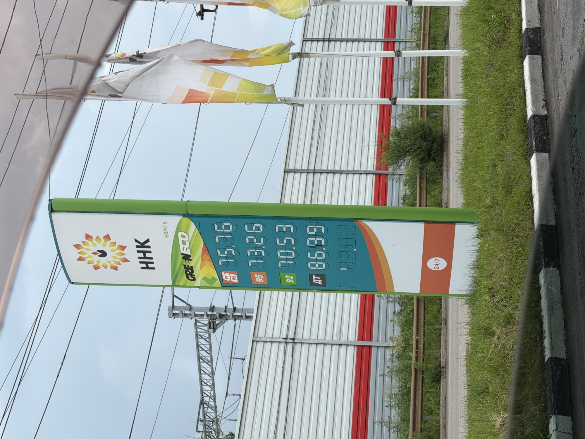 The image shows a gas station price sign with various fuel prices displayed. The sign is green and white with colorful accents. The prices listed are for "GE 95" at 75.76, "95" at 73.26, "92" at 70.53, and "AT" at 86.99. There is also a section below displaying "00.00". Above the prices, the brand "HHK" and "GREEN ECO" are visible, along with the text "ЕВРО 5". The sign has a "24/7" symbol. In the background, there is a metal fence, grass, a road, and electrical wires with a train track structure. Several flags are also visible on poles to the right of the sign. The overall scene suggests a roadside location, likely a gas station. The provided context indicates the location is Khabarovsk, Russia.