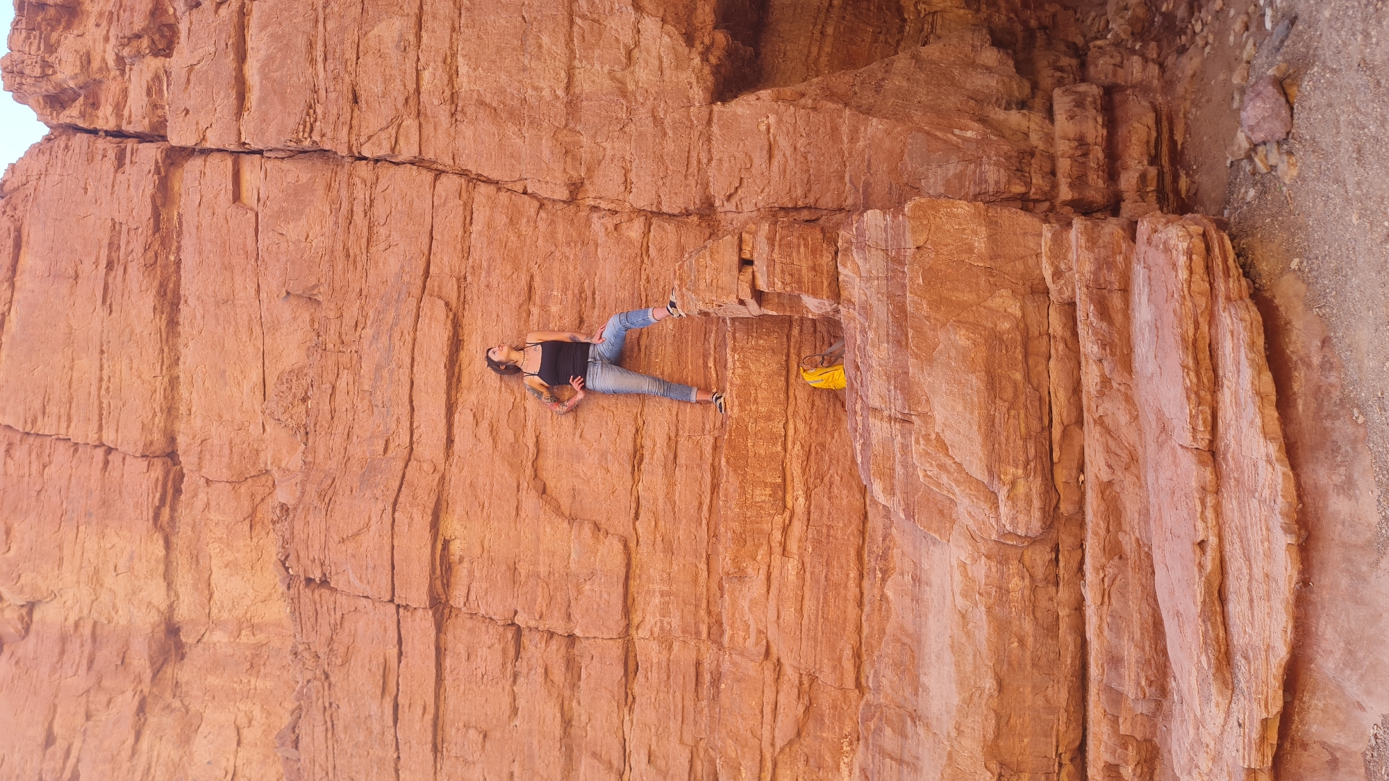 A woman wearing a black tank top and jeans reclines on a red sandstone rock formation, possibly in or near the city of Hevel Eilot Regional Council, Israel. A yellow backpack is partially visible next to her, lodged between the rocks. The backdrop is a textured red rock face.