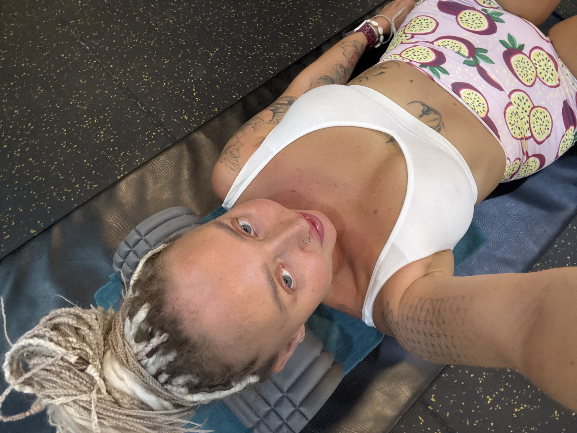A woman with blonde dreadlocks and tattoos is lying on a gym mat in a gym. She is wearing a white sports bra and pink shorts with a passion fruit print. The gym floor is black with yellow speckles. The image is taken from a high angle, looking down at the woman. The location is stated as Bac Nha Trang Ward, Vietnam.