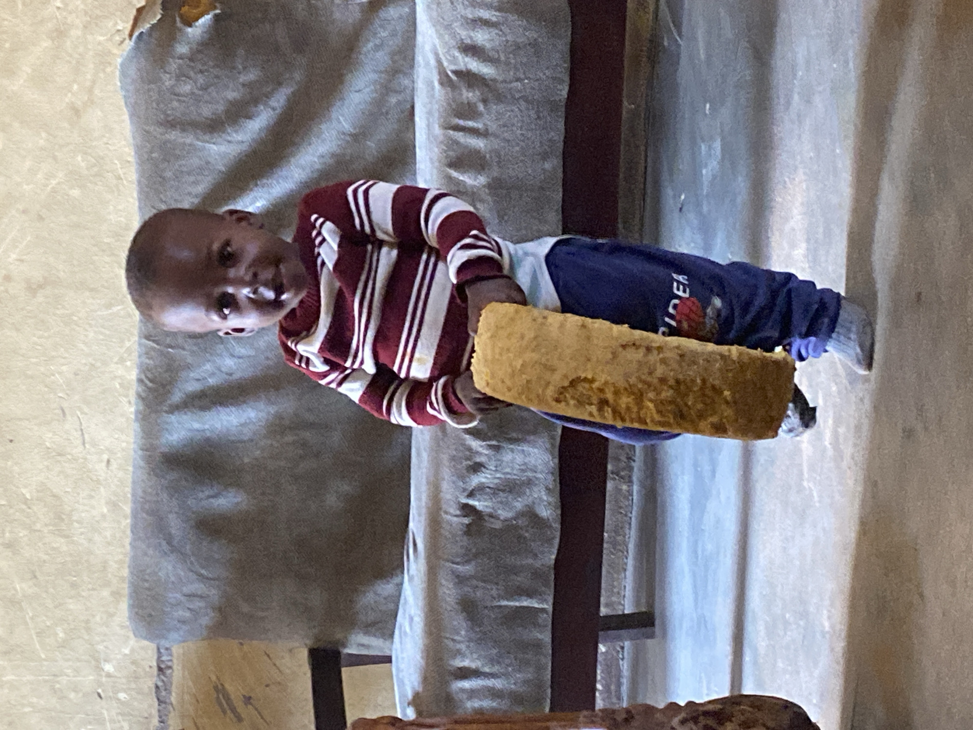 A young child, likely a boy given the clothing and short hair, stands in an indoor setting holding a large, elongated piece of fried dough or bread. The child wears a striped long-sleeved shirt in red and white and dark blue pants with a red graphic and white text on the left leg. Light grey socks cover the child's feet, and one foot appears to be wearing a dark shoe. The background features a neutral-colored couch with a textured surface and a light-colored, slightly worn wall. The floor is a light grey, concrete-like material. The lighting suggests an indoor environment, possibly in a home or communal space. The image captures a moment of the child holding the food item, looking towards the camera with a slight smile. The scene appears to be located in Panyam, Nigeria, based on the provided context.