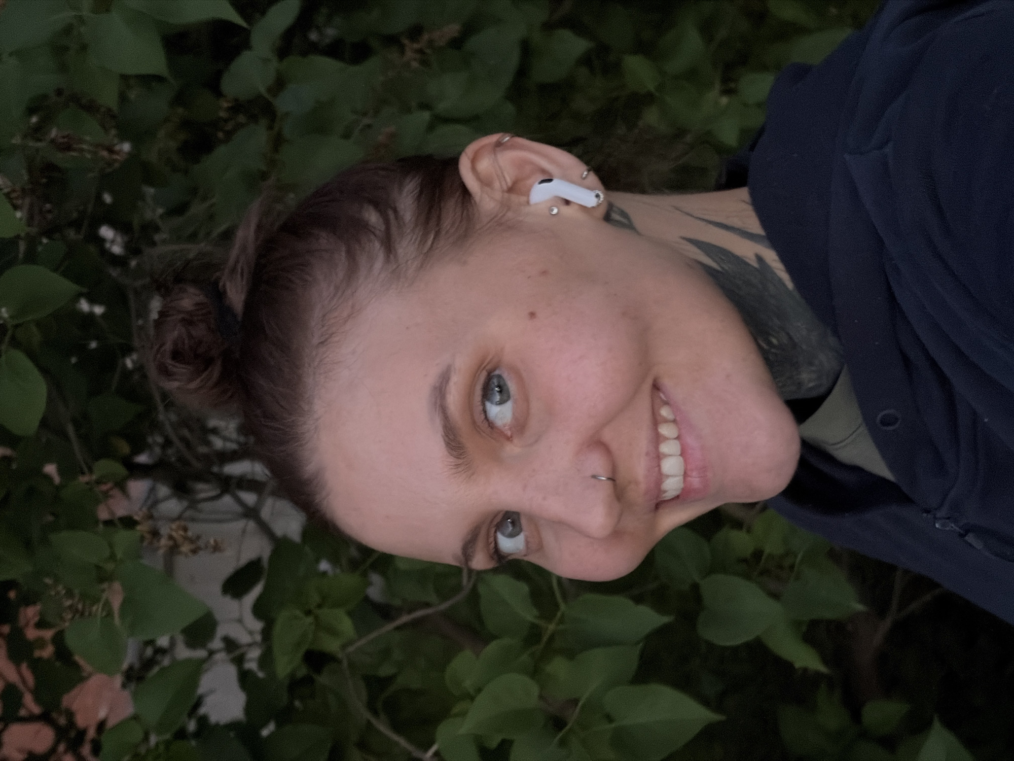 A medium close-up portrait shows a young woman with her hair in a bun, wearing a dark blue hoodie and an olive green shirt. She has a nose ring and multiple earrings, including white wireless earbuds in her right ear. A tattoo is visible on her neck. The woman is smiling and looking towards the camera. She is positioned in front of a backdrop of green foliage, which appears to be outdoors. The scene is dimly lit, suggesting it may be dusk or nighttime. There is no identifiable urban or city landscape visible in the image.