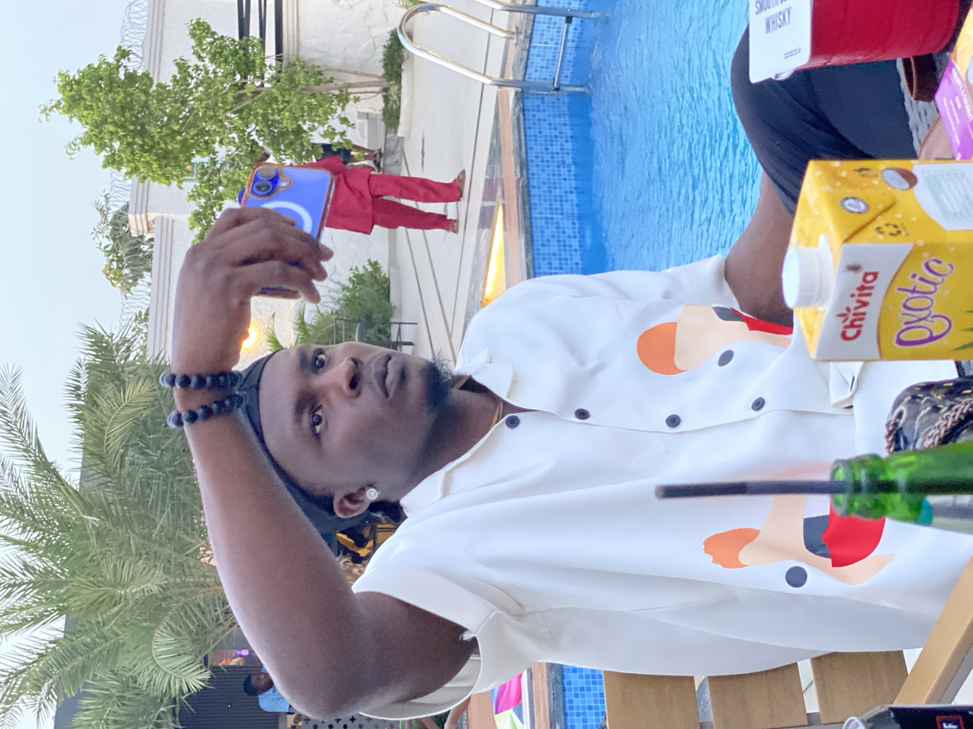 A man is seated outdoors next to a swimming pool, holding up a blue smartphone to take a photo. He is wearing a white shirt with cartoonish human figures printed on it, and a beaded bracelet. Next to him is a carton of Chivita Exortic juice and a green bottle. In the background, another individual is wearing a red outfit, and the swimming pool is visible. The location is identified as Kubwa, Nigeria.