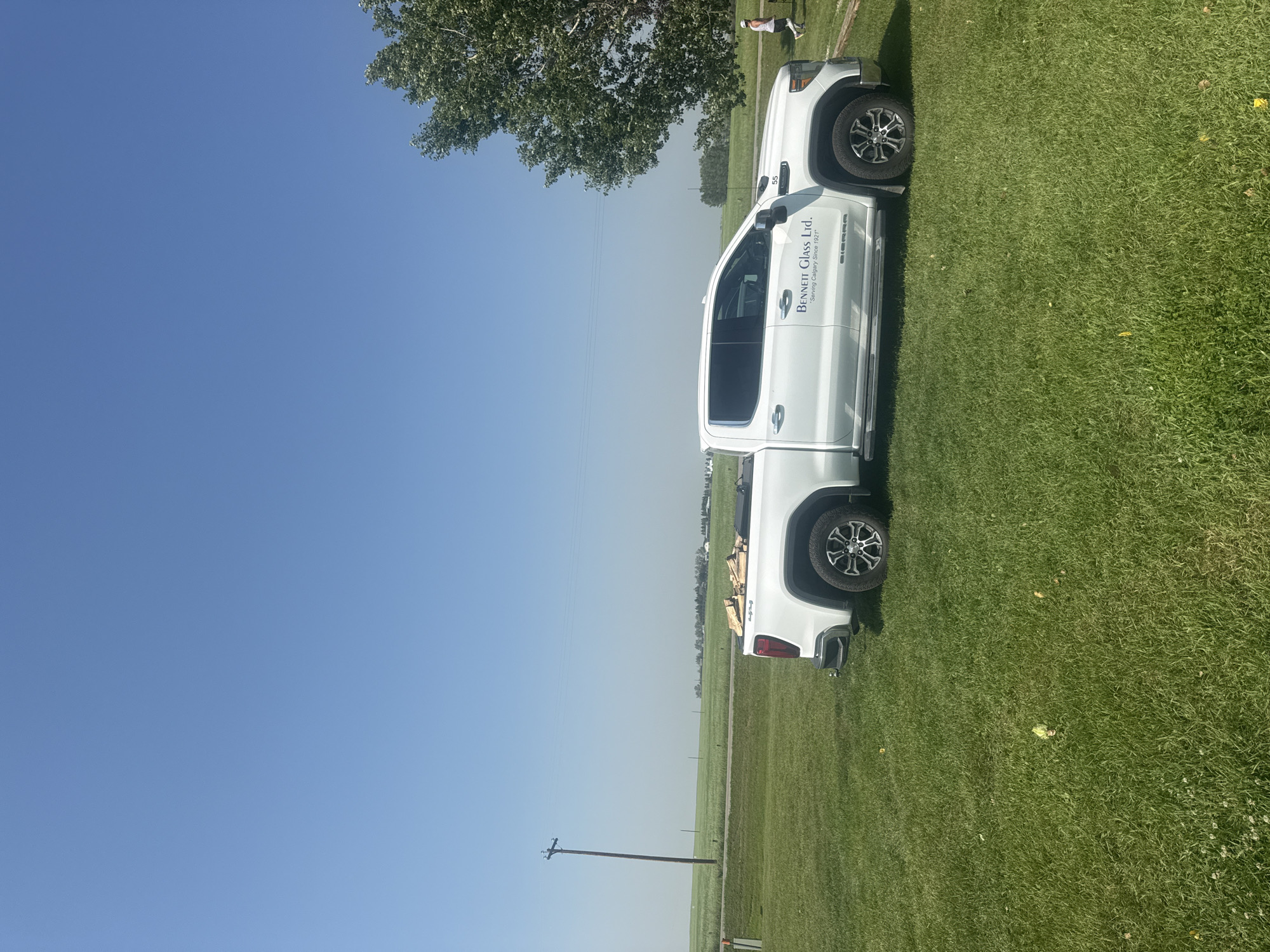 In a rural setting in Alberta, Canada, a white GMC Sierra pickup truck is parked on a grassy field under a clear blue sky. The truck bed contains several pieces of firewood. A large green tree is visible to the right of the truck, and in the distance, a person can be seen walking. The landscape appears to be farmland, with fields stretching to the horizon and a solitary power pole standing in the middle distance. The scene is illuminated by bright sunlight.