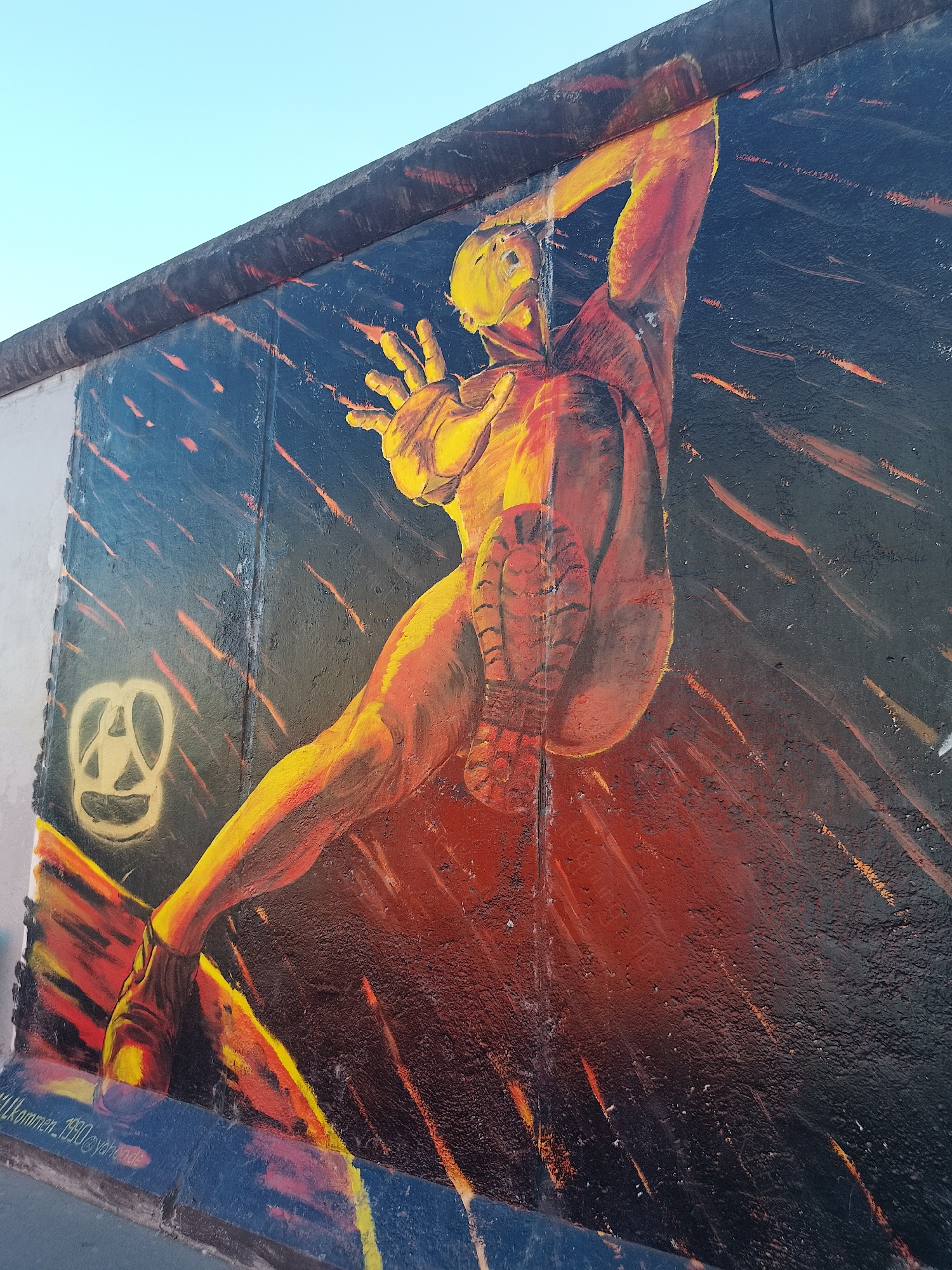 The image is a section of the Berlin Wall, now serving as an art canvas. On the wall is a mural of a figure, painted in shades of orange, red, and yellow, seemingly in motion. The figure is nude and appears to be climbing or reaching upwards, one arm extended and hand open as if grasping something. The other arm is raised higher. The lower body of the figure is oriented toward the viewer, showing the sole of one shoe.
The background of the mural transitions from a dark blue at the top to red and black lower down, with thin streaks of orange throughout. To the left of the figure is a yellow circular symbol. At the base of the wall, there is some visible graffiti and possible signatures. The top of the wall has a dark gray ledge. The sky above the wall is light blue.
The location is the East Side Gallery in Berlin, Germany, the longest remaining section of the Berlin Wall and now an open-air art gallery.